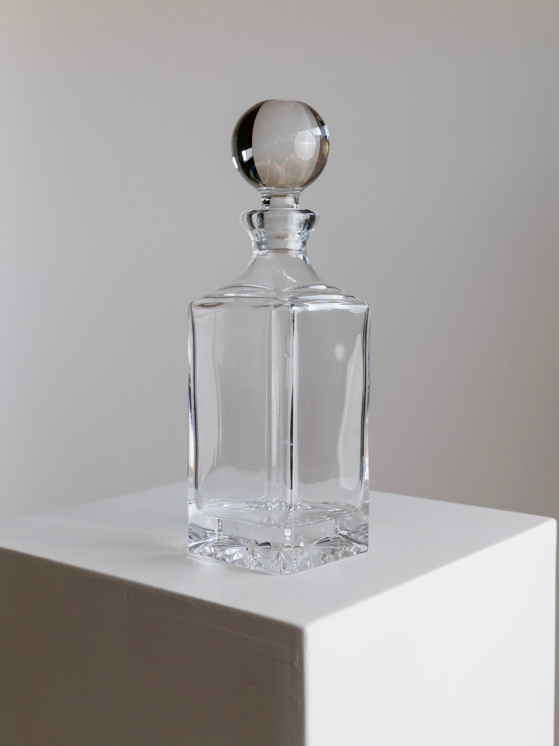 The Murano Glass Carafe 70s by Bottega Jacobs, featuring a clear rectangular design and a spherical stopper, is displayed on a white pedestal against a neutral backdrop. The empty decanter captures and reflects light, showcasing its smooth and elegant craftsmanship.