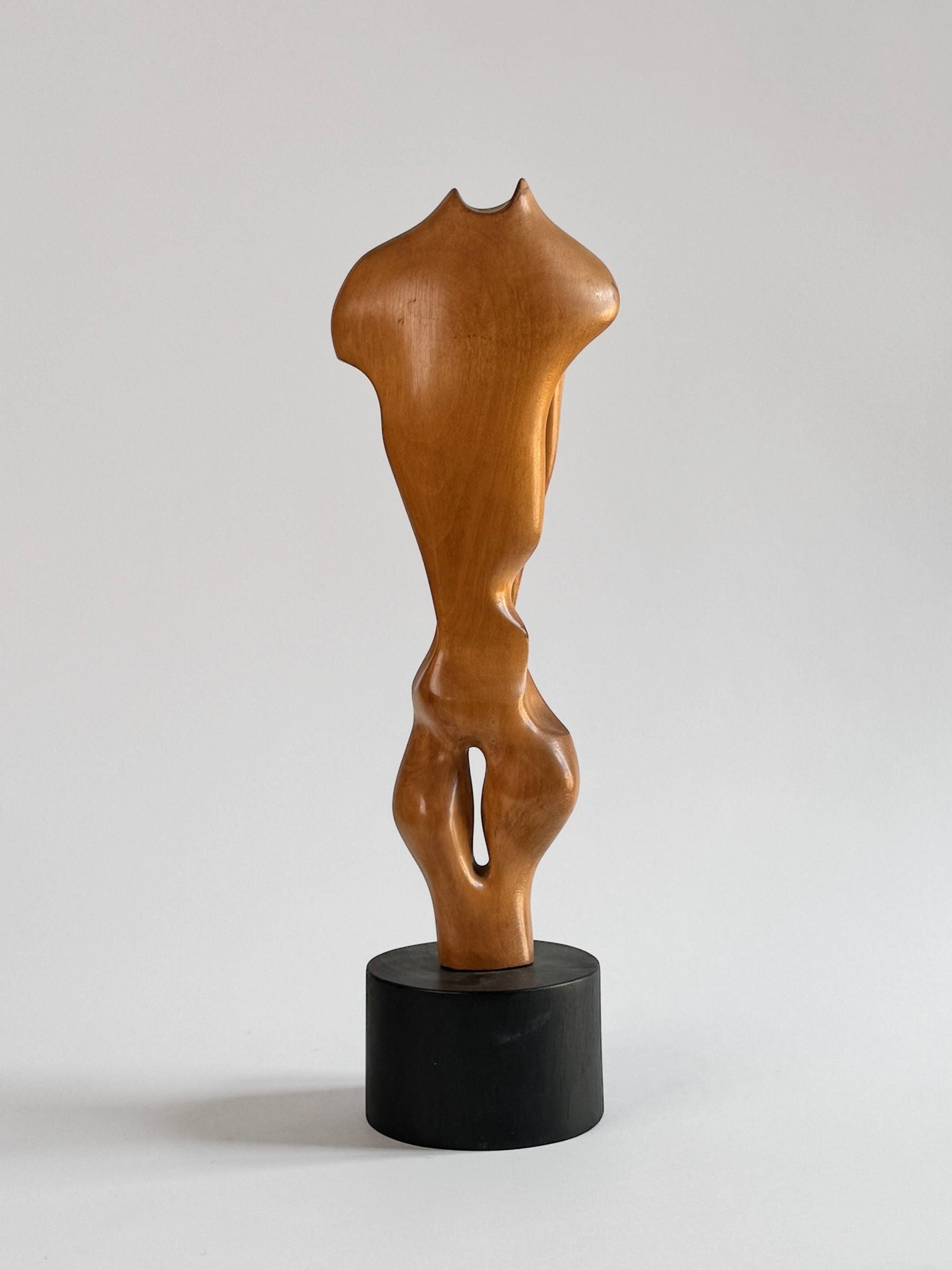 Introducing the Wood Sculpture by Pierre Dedieu, a masterpiece by Figuresdesign. This varnished walnut wood sculpture is elegantly presented on a cylindrical black base. Featuring smooth, curved lines, it subtly evokes the form of a torso while maintaining an intentionally ambiguous and stylized design that encourages multiple interpretations. The plain light grey background enhances its striking presence.