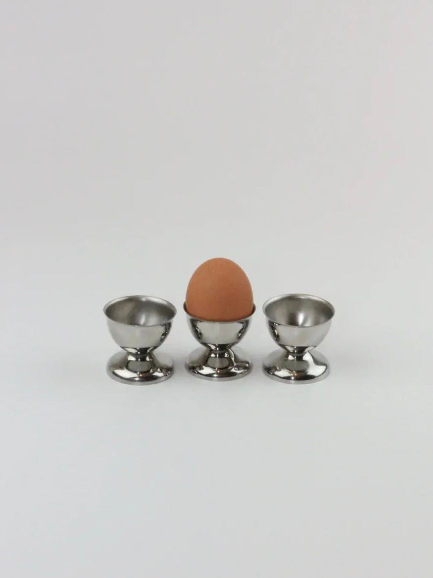 A single brown egg rests in the middle of three shiny stainless steel "Six Inox Egg Cups" from Boga Avante Shop, neatly arranged in a row on a plain white background. One egg cup holds the egg, while the other two remain empty. These elegant breakfast pieces from Boga Avante Shop showcase both durability and strength.