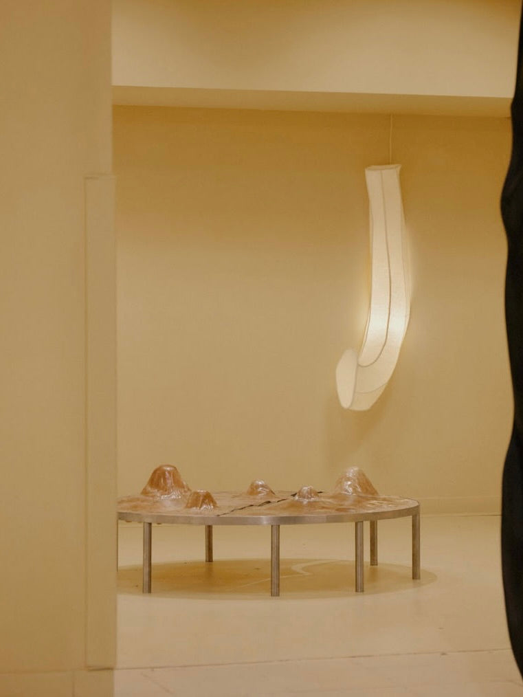 A minimalist room with beige walls showcases Panorammma's Phantom: Rawhide table, topped with a sculpture resembling small hills in transparent material. Nearby, an elongated light fixture exudes a phantasmagoric quality from the ceiling.
