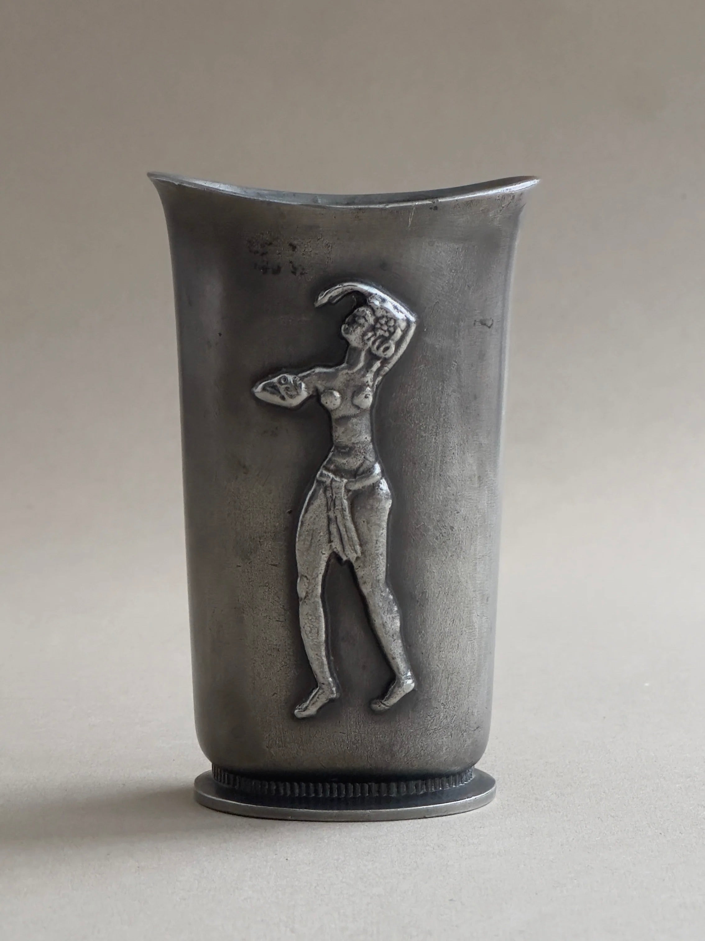 The Galerie Storm Art Deco vase in Cast Pewter, GAB, is a tall silver cylindrical piece featuring a high-relief woman in draped attire. It elegantly combines Art Deco style with Swedish Grace against a neutral gray backdrop.