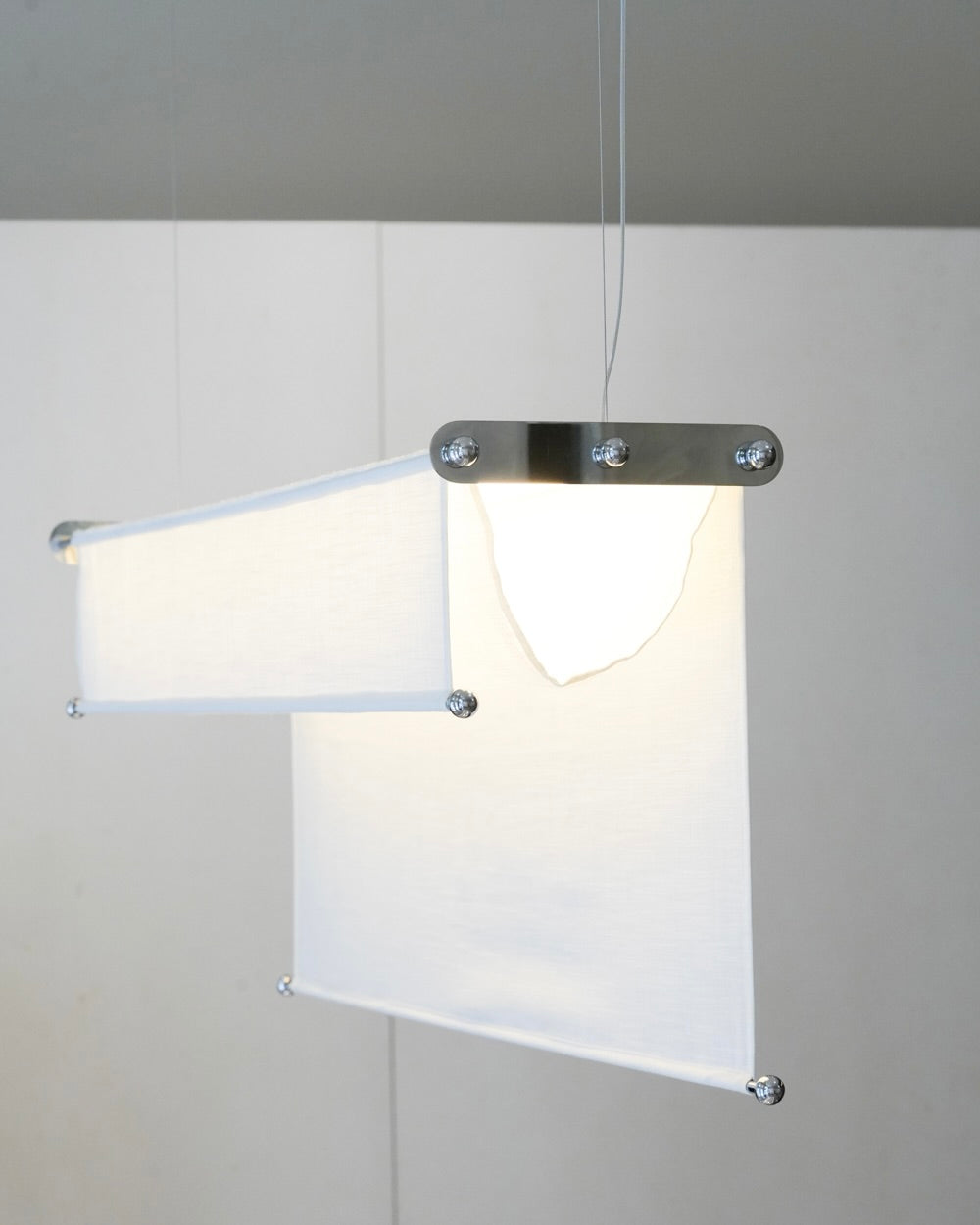 The Vento Pendant by Caia Leifsdotter features two adjustable rectangular fabric panels suspended from a stainless steel base with thin wires, offering a modern, organic look.