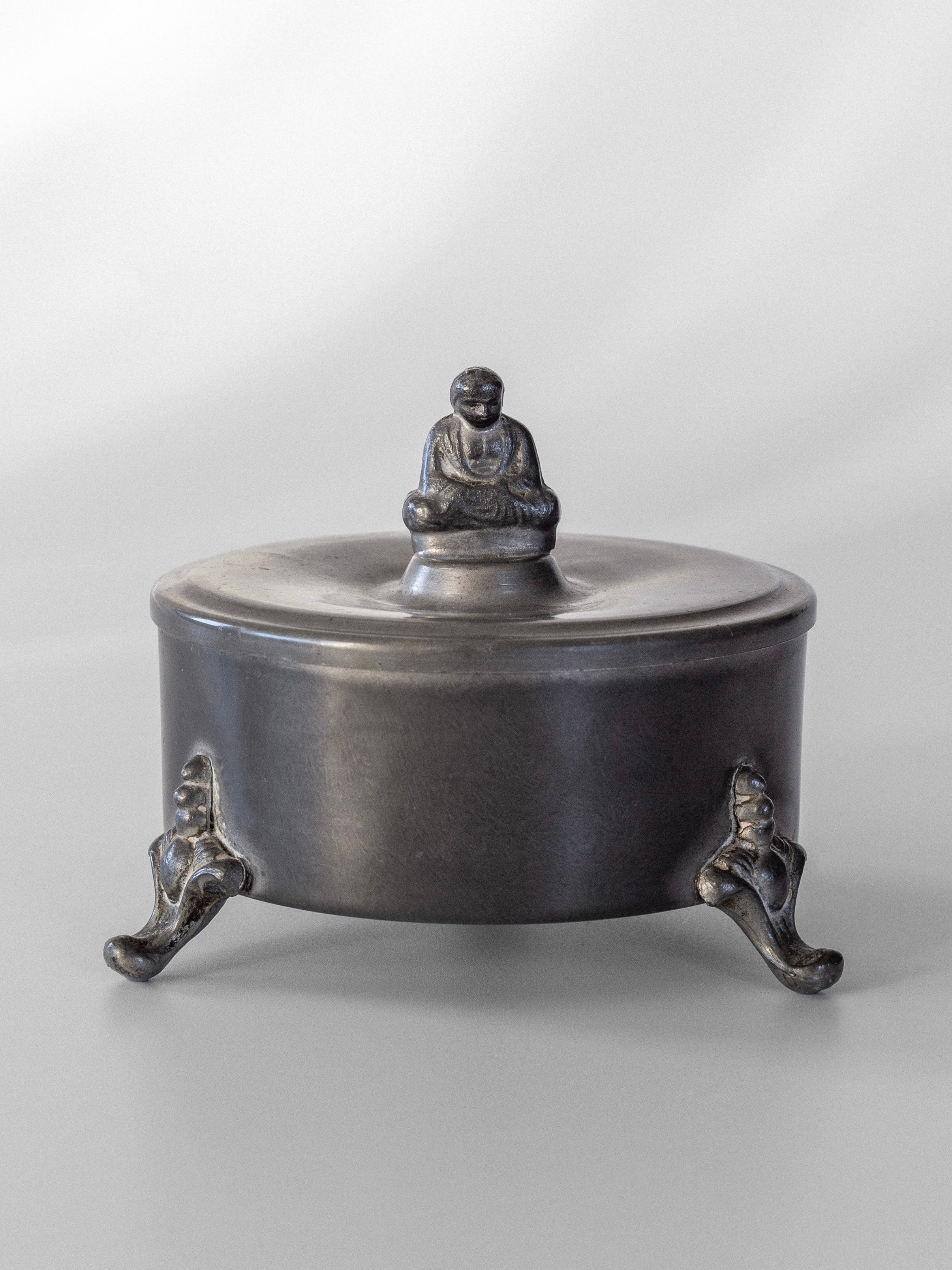 A round Art Deco pewter box with a lid showcases a small seated Buddha figure on top. Designed by Spigel, it is supported by three ornate legs, providing an elevated look against a neutral gray background.