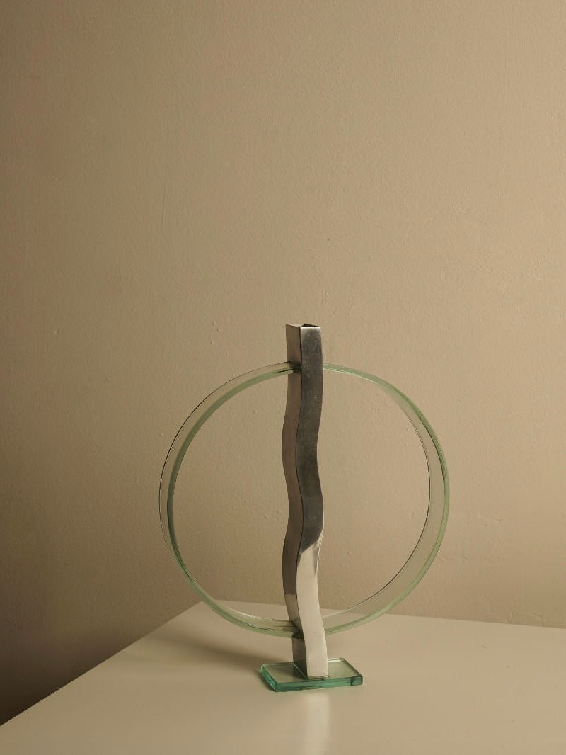 A minimalist metallic and glass sculpture featuring a circular glass frame intersected by a wavy metallic column, placed on a light-colored tabletop accessorized with the Wavy Small Vase by Les Objets Amsterdam, against the backdrop of a beige wall. This piece exudes Provençal Prestige and is perfect for premium homeware collections.