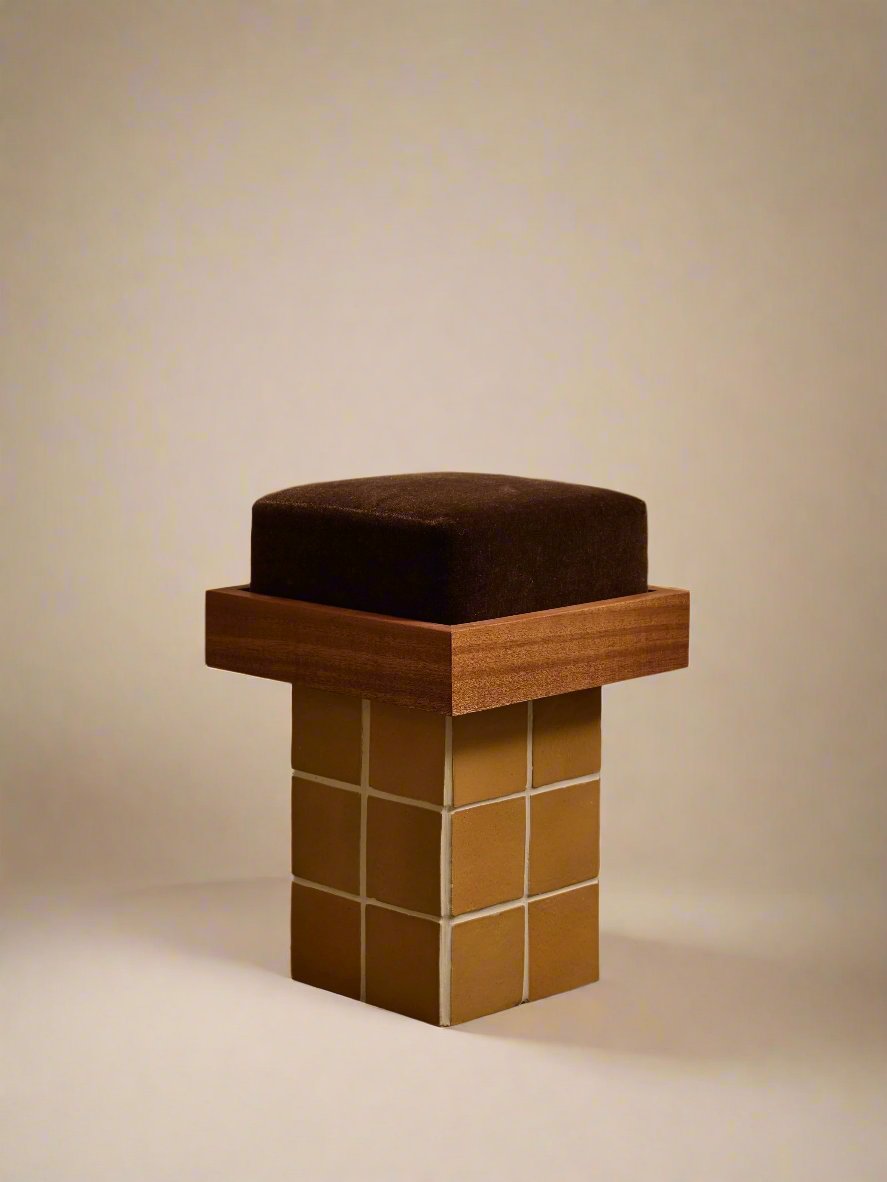 The Jura Stool by Ah Um Design Studio is a modern, minimalistic chair featuring a brown upholstered mohair cushion, a wooden platform of mahogany with an oil finish, and a base made of yellow square tiles with white grout lines. The stool sits on a concrete floor against beige curtains in the background.