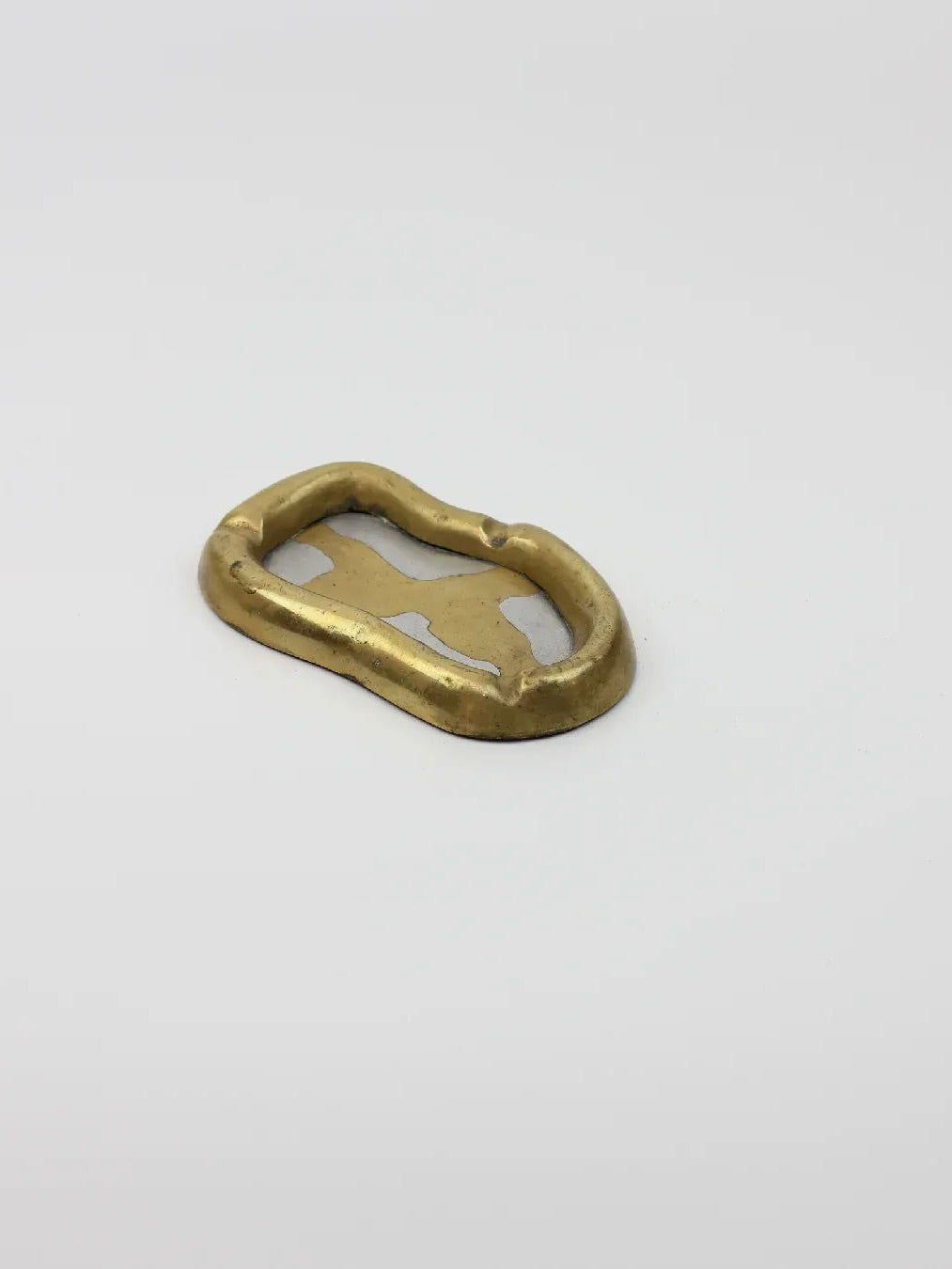 A gold-toned Brutalist Ash Tray from the Boga Avante Shop rests on a plain white surface. Its irregular shape, rustic brass finish, and slightly uneven edges embody a brutalist style with an open center.