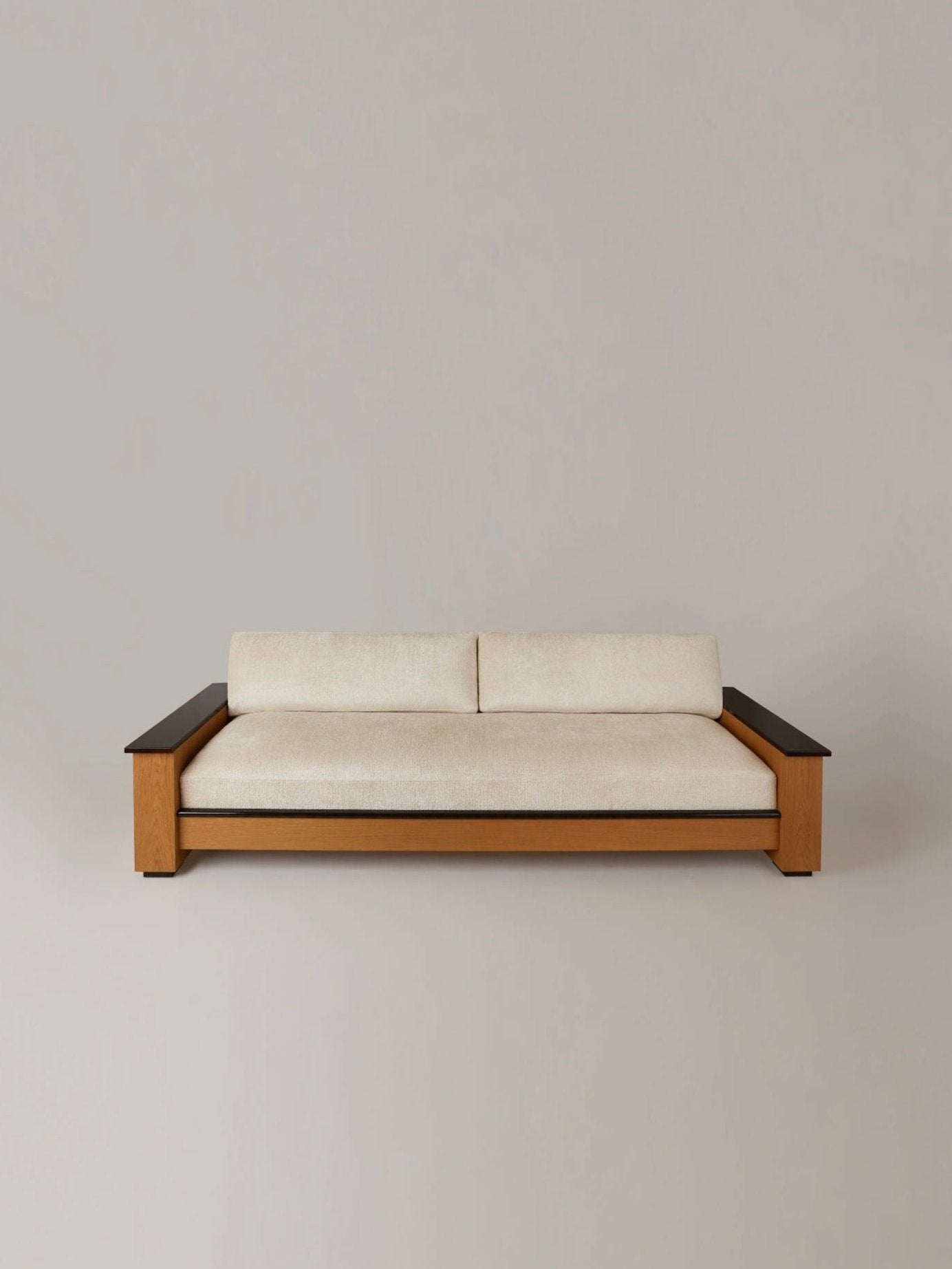 The "Le Colonel" Sofa by HAUVETTE & MADANI, a minimalist beige piece with a wooden shell and armrests, sits against a neutral light gray background. The modern design boasts clean, straight lines and includes optional cushions for enhanced comfort.