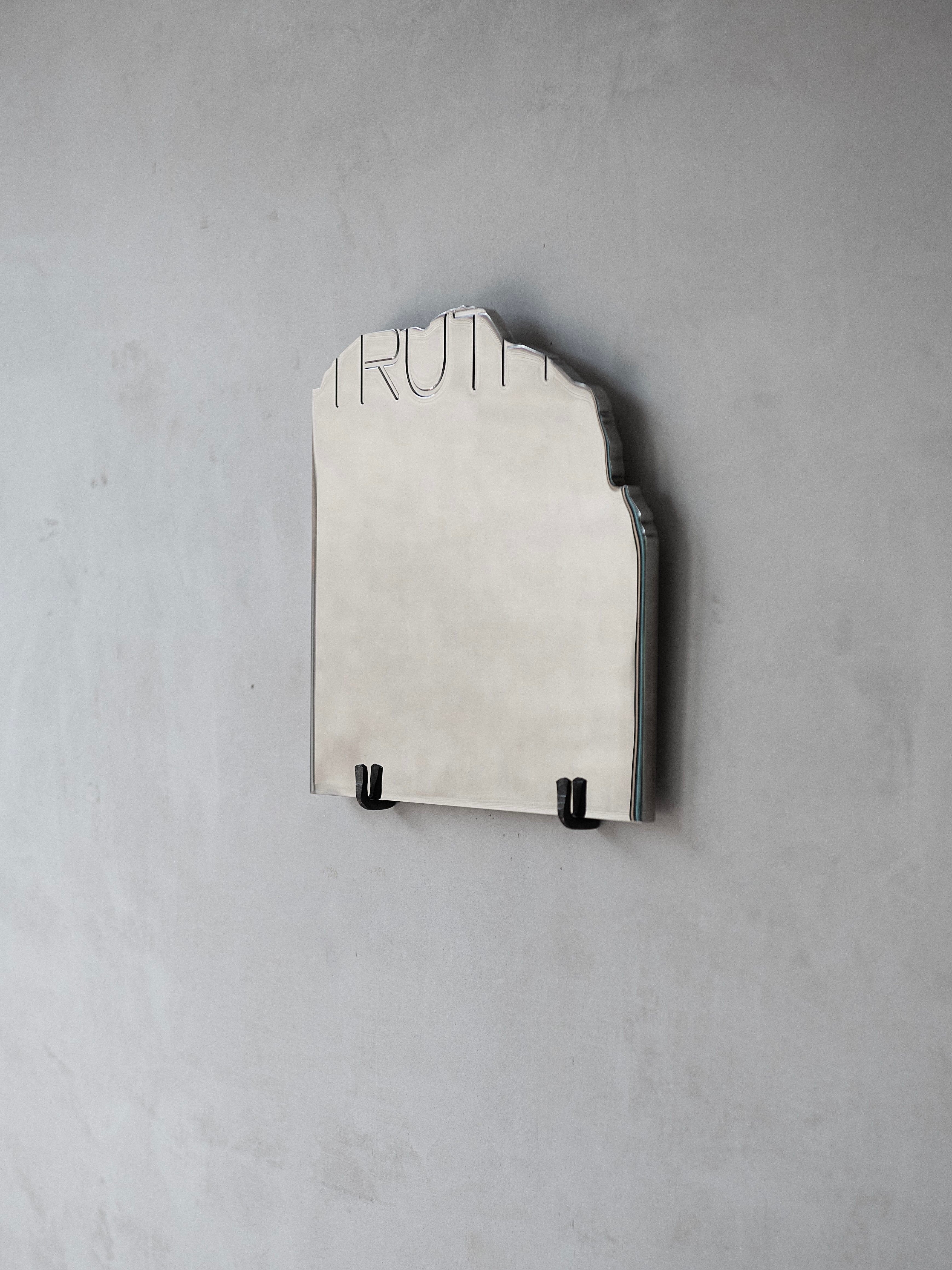 A "THE TRUTH" Mirror by COSEINCORSO, uniquely shaped with "TRUTH" etched along the top edge, is mounted on a bare gray wall, reminiscent of an ancient epigraph. Supported by two small black brackets, this mirror enhances its minimalist and modern aesthetic.