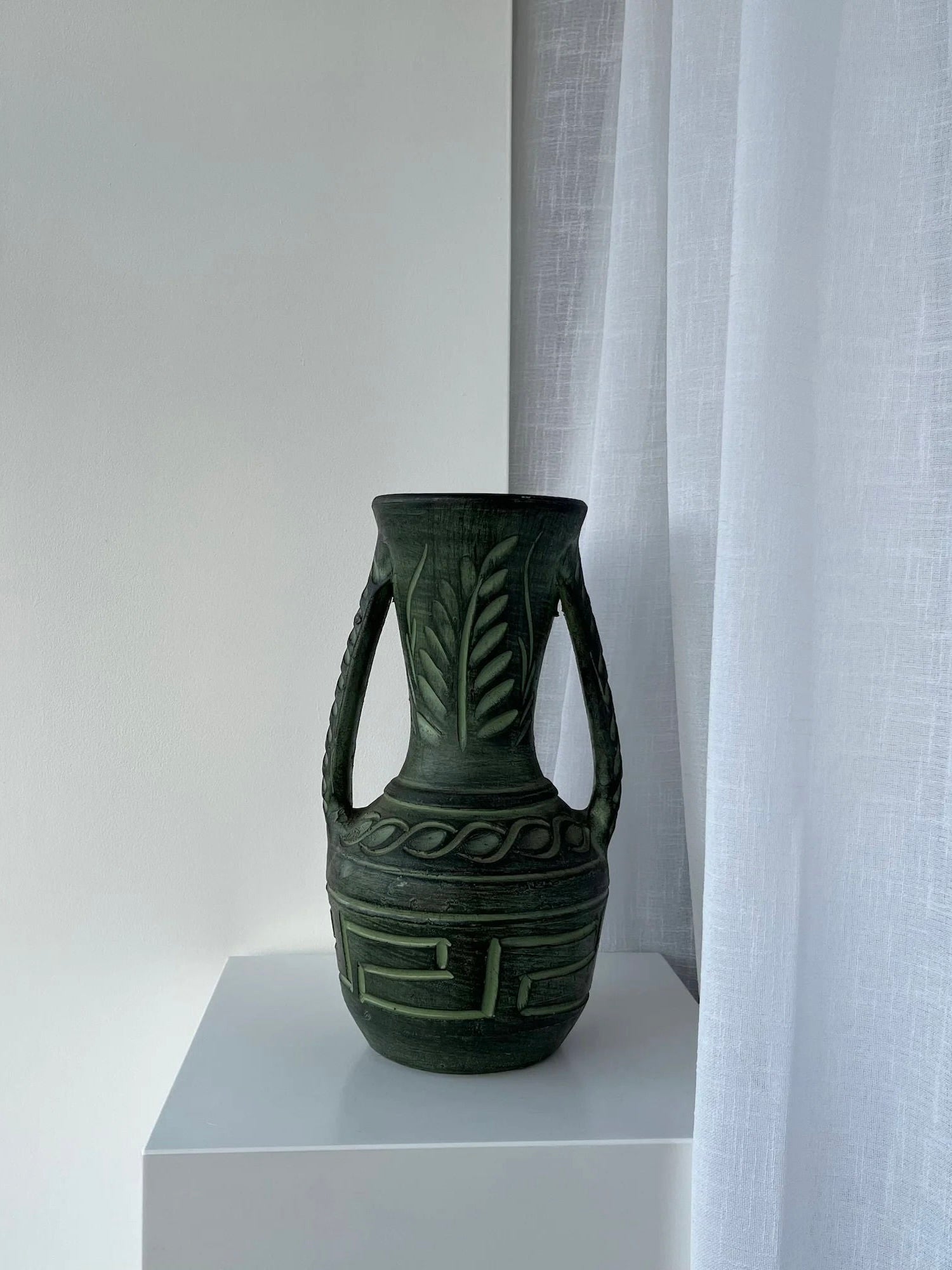 The European Verdigris Large Vase from Aesthete Label stands on a white pedestal, showcasing its stunning dark green ceramic with intricate carvings. The middle section features an elegant leaf pattern, while the handles boast a sophisticated braided design. This large statement piece is finished with a matt verdigris finish. The white wall and sheer curtain in the background provide a perfect backdrop for this artistic creation.