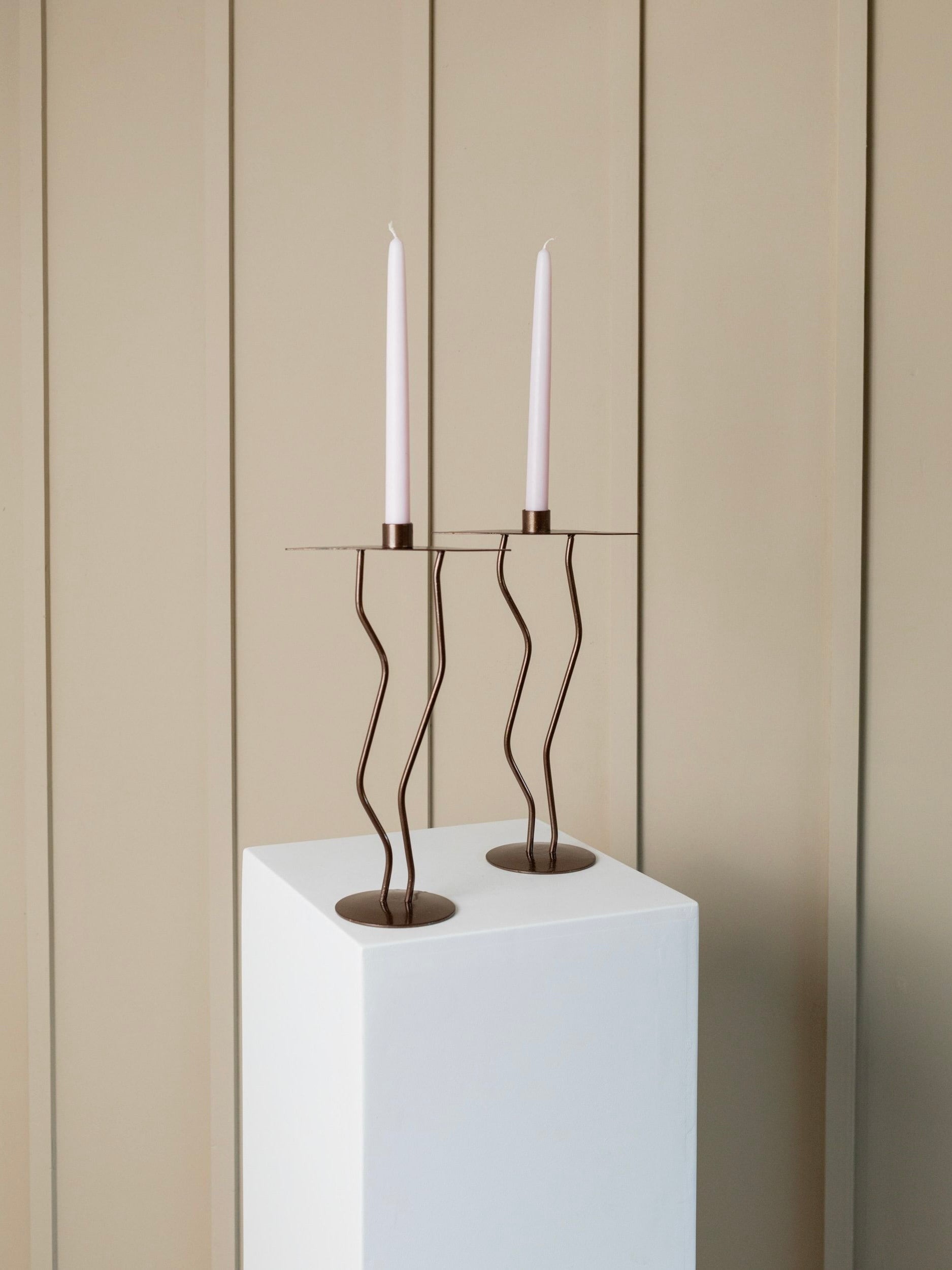A pair of slender, tall, pink candles are elegantly displayed on the Bottega Jacobs' "Two Metal Candlesticks Memphis Style" atop a white pedestal. These bronze-painted holders enhance the beige paneling in the background, culminating in a minimalist and chic setting with a subtle touch of Memphis style flair.