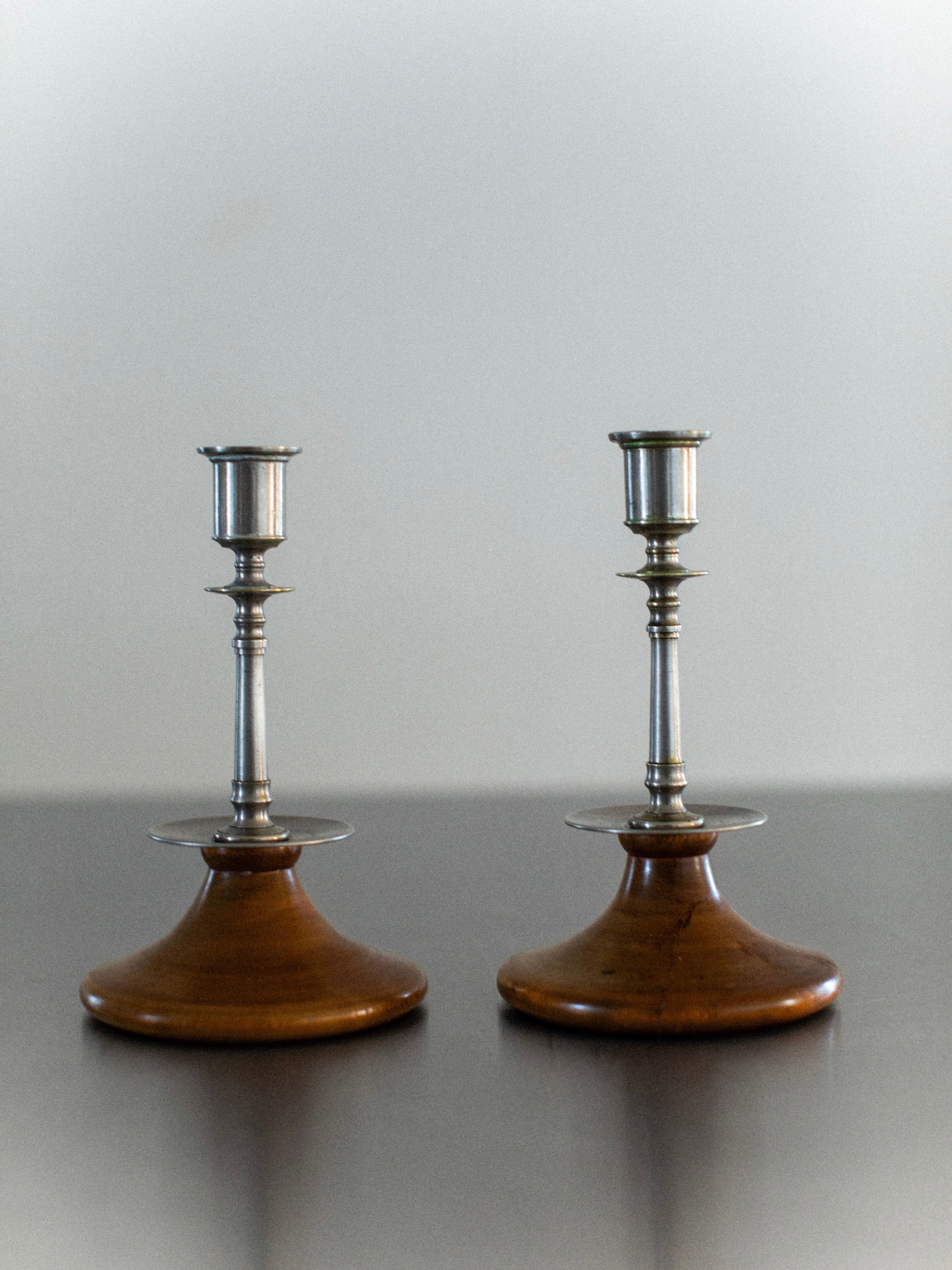The Steel & Wood Candleholders from Out For Lunch exude rustic charm as they stand side by side on a smooth, dark surface, while the softly blurred background highlights their elegant design.