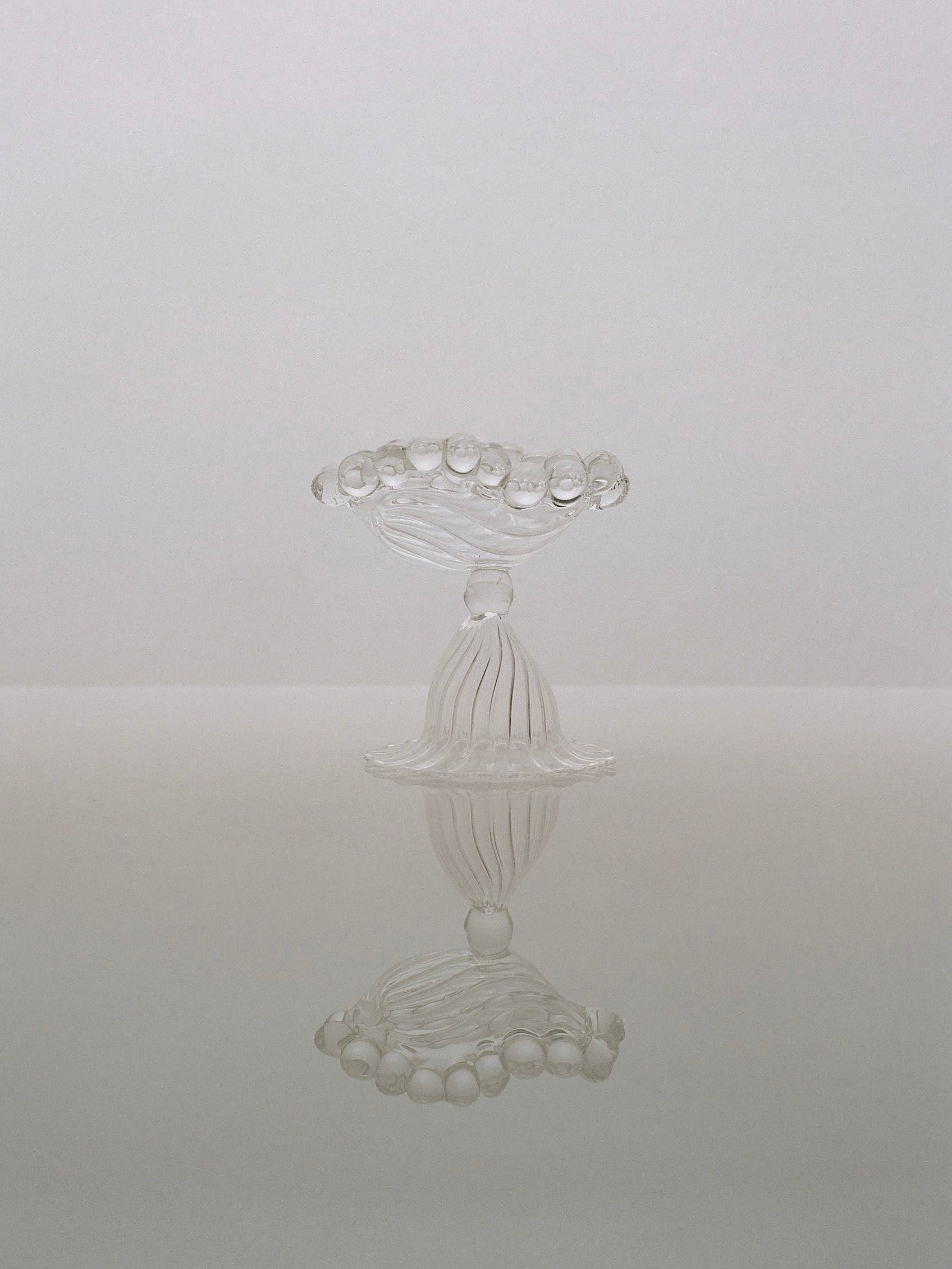 The Perséphone Ashtray by Justine Menard, with its clear, intricately designed glass and scalloped edge, resembles a hand-blown table sculpture. It is placed against a neutral, reflective background, giving an impression of symmetry and elegance.