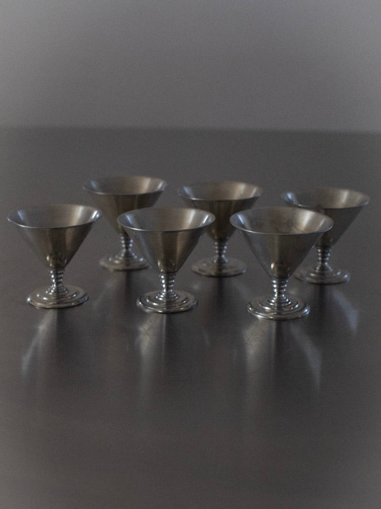 A set of "Six Ice Cream Cups" by Out For Lunch, crafted from silver metal and echoing the charm of vintage martini glasses, are arranged in two rows of three on a reflective surface. Featuring a minimalist design with an elegant, tapering shape, the softly blurred background directs all attention to these exquisite cups.