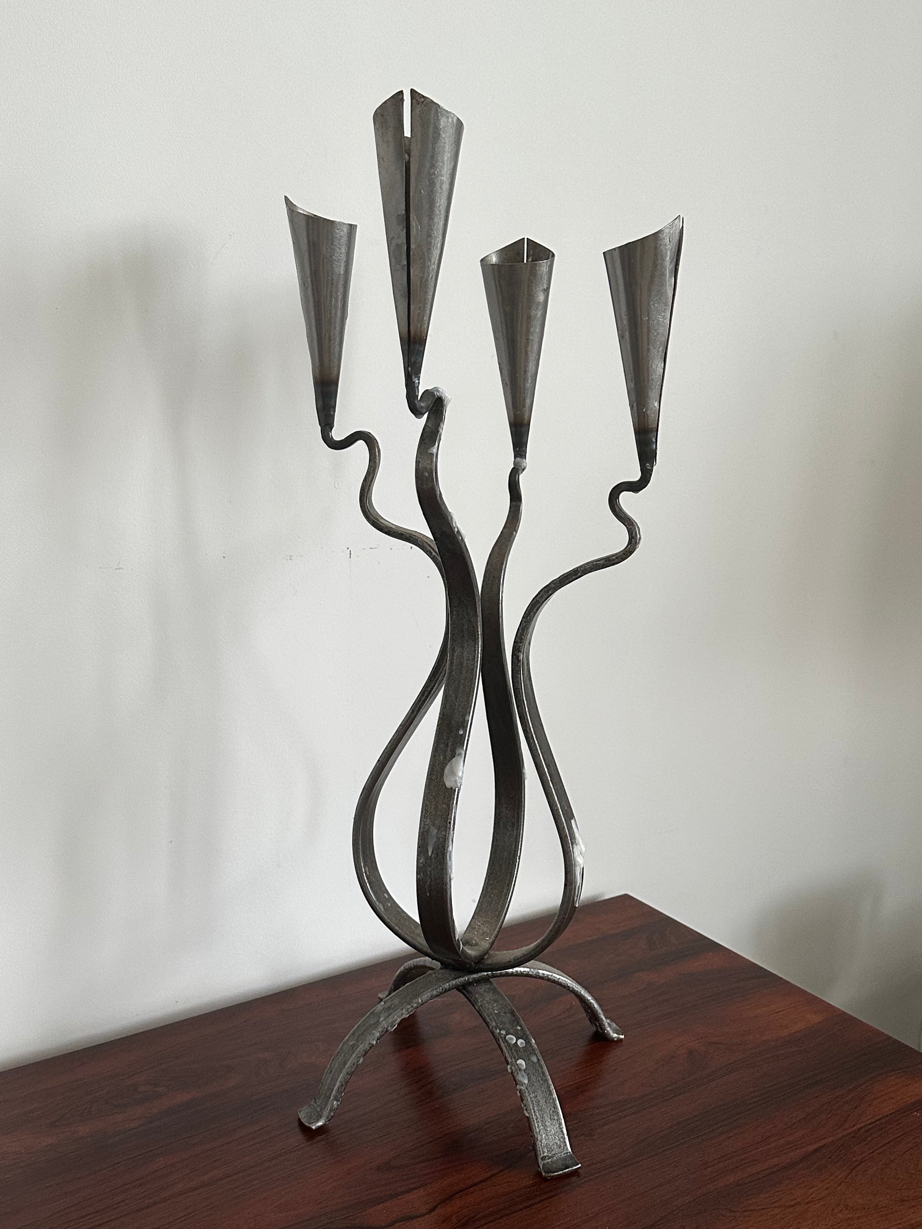 The Aesthete Label Post Modern Steel Large Candle Holder features three twisted arms with cone-shaped holders, creating a striking accent for modern decor when displayed on a dark wooden surface against a white wall.