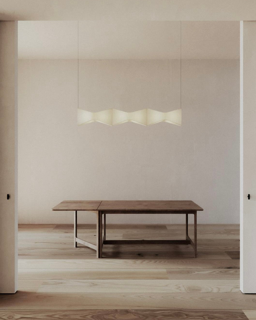 A minimalist interior with a wooden table and a modern "Arête" Pendant Horizontal Line by Asca Studio hanging above. Light wooden flooring, neutral walls, and wheat-colored linen accents enhance the serene atmosphere and sculptural sensibility.