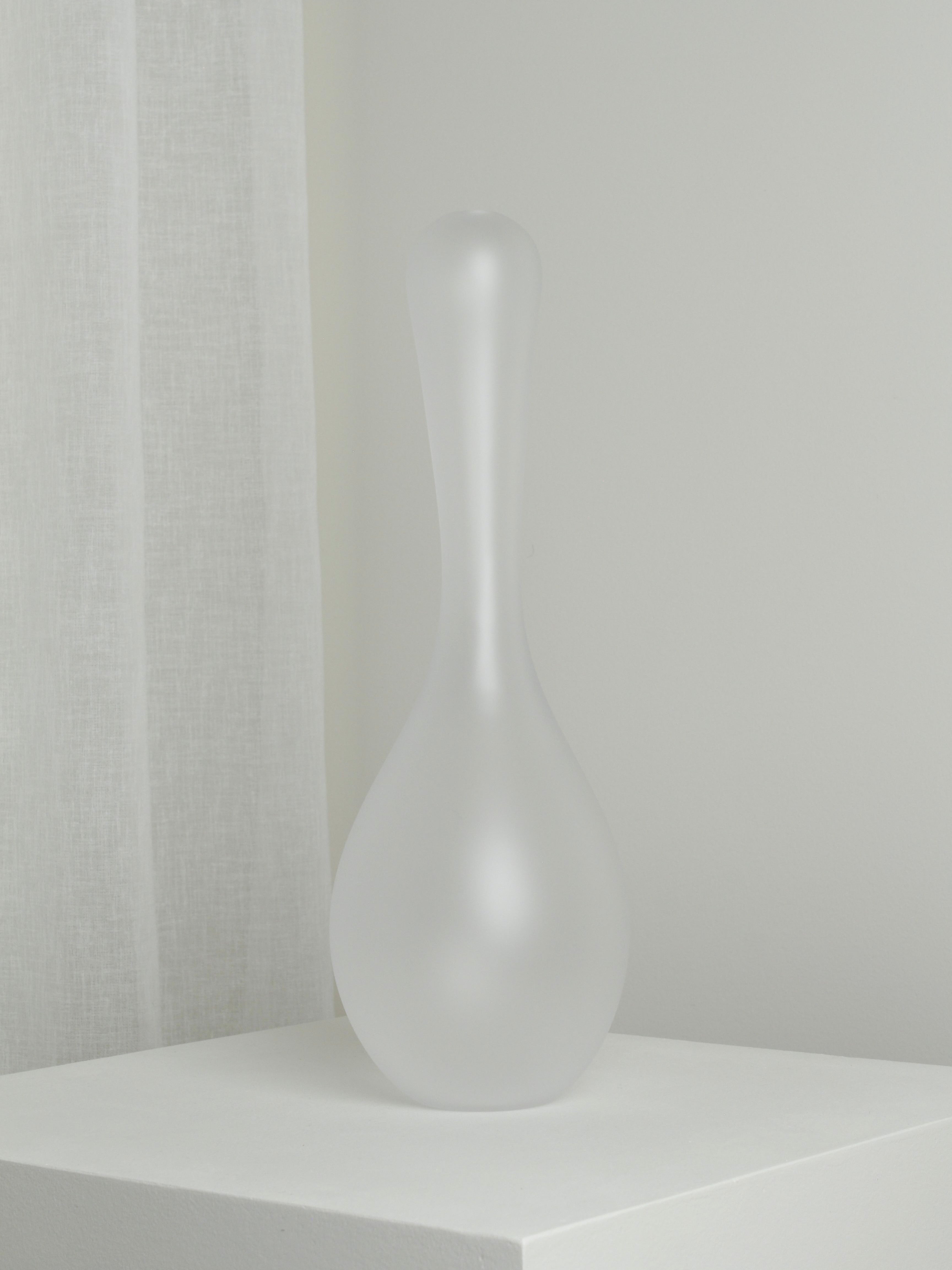 The Verre d'Onge XLarge Vase in Ghost, featuring a narrow neck and bulbous base, is placed on a white pedestal. A light gray wall and sheer white curtain create a serene, minimalist backdrop, giving the scene an almost ghost-like presence.