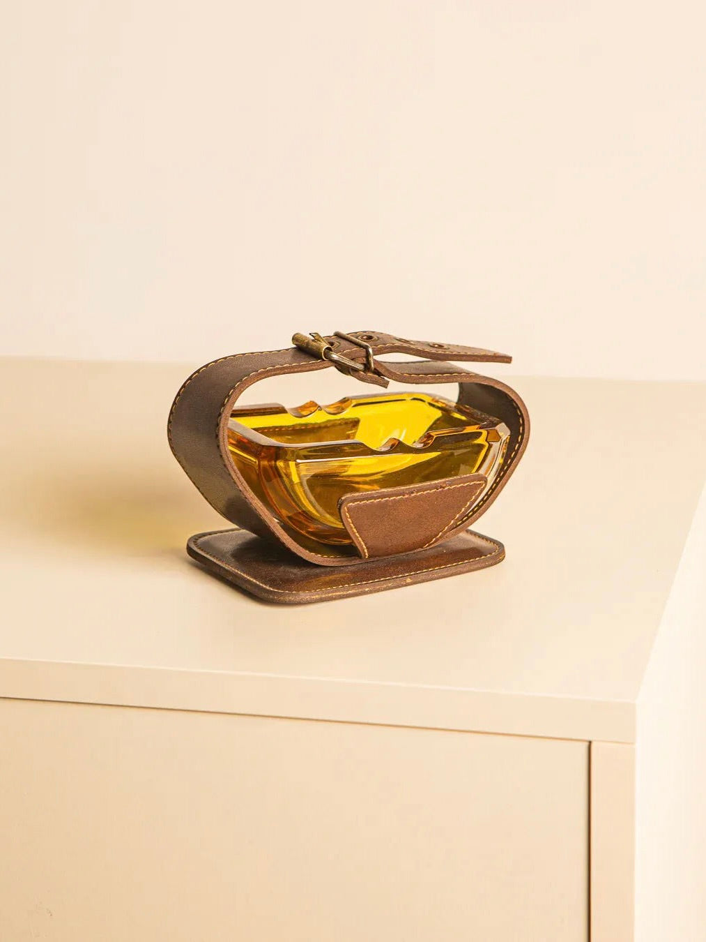 On a white surface rests an Italian Ashtray from the 60s by Treaptyque, its vintage design highlighted by a leather wrap. The ashtray, filled with amber tones, mimics the shape of an apple with a decorative flair. A zipper detail enhances the leather wrap, contributing to its distinctive charm.