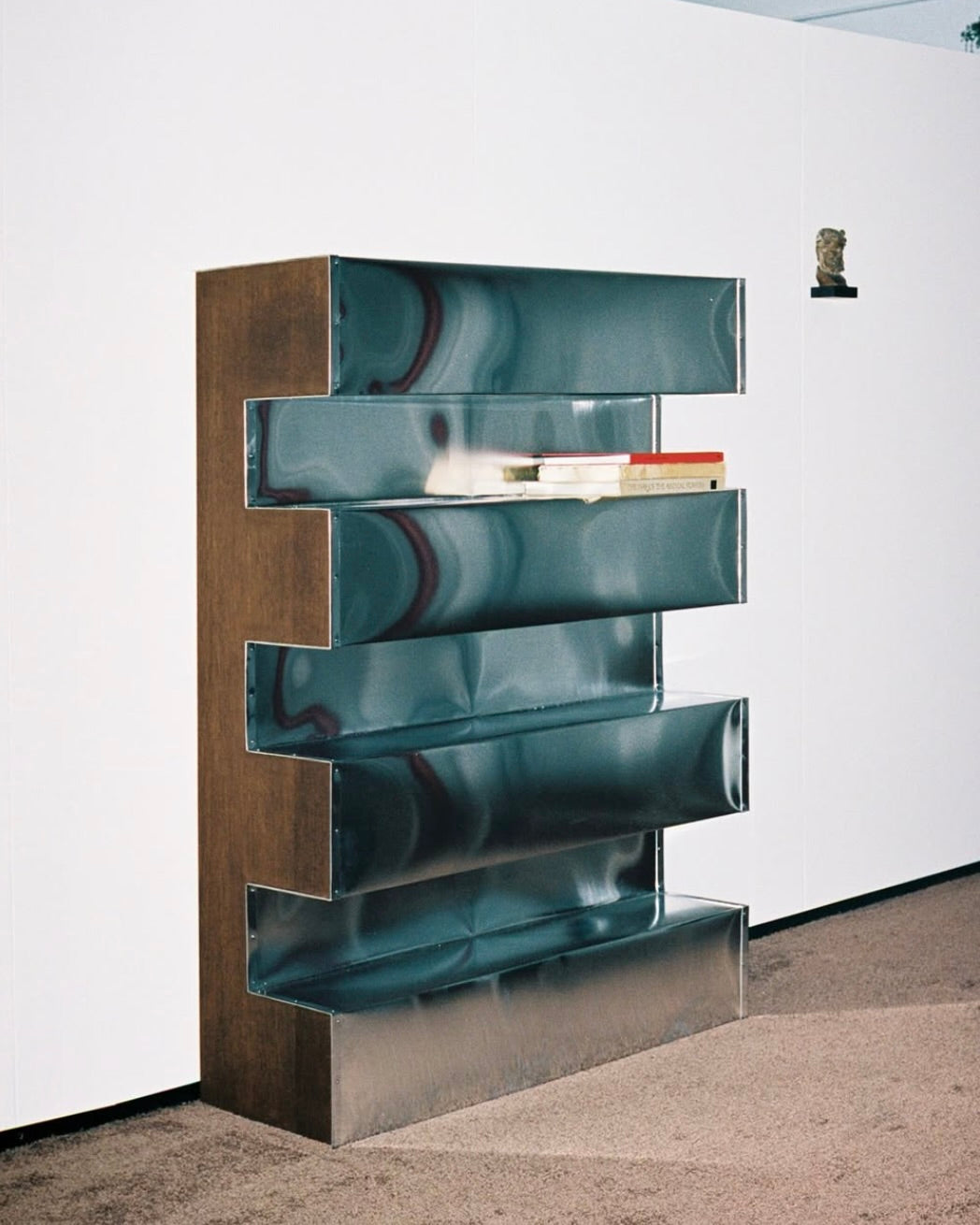 The Aluminum Bookshelf by Studio HAOS features five staggered shelves in geometric sheet aluminum. Books and magazines rest on one shelf, while a small sculpture is displayed on a nearby wall-mounted shelf.