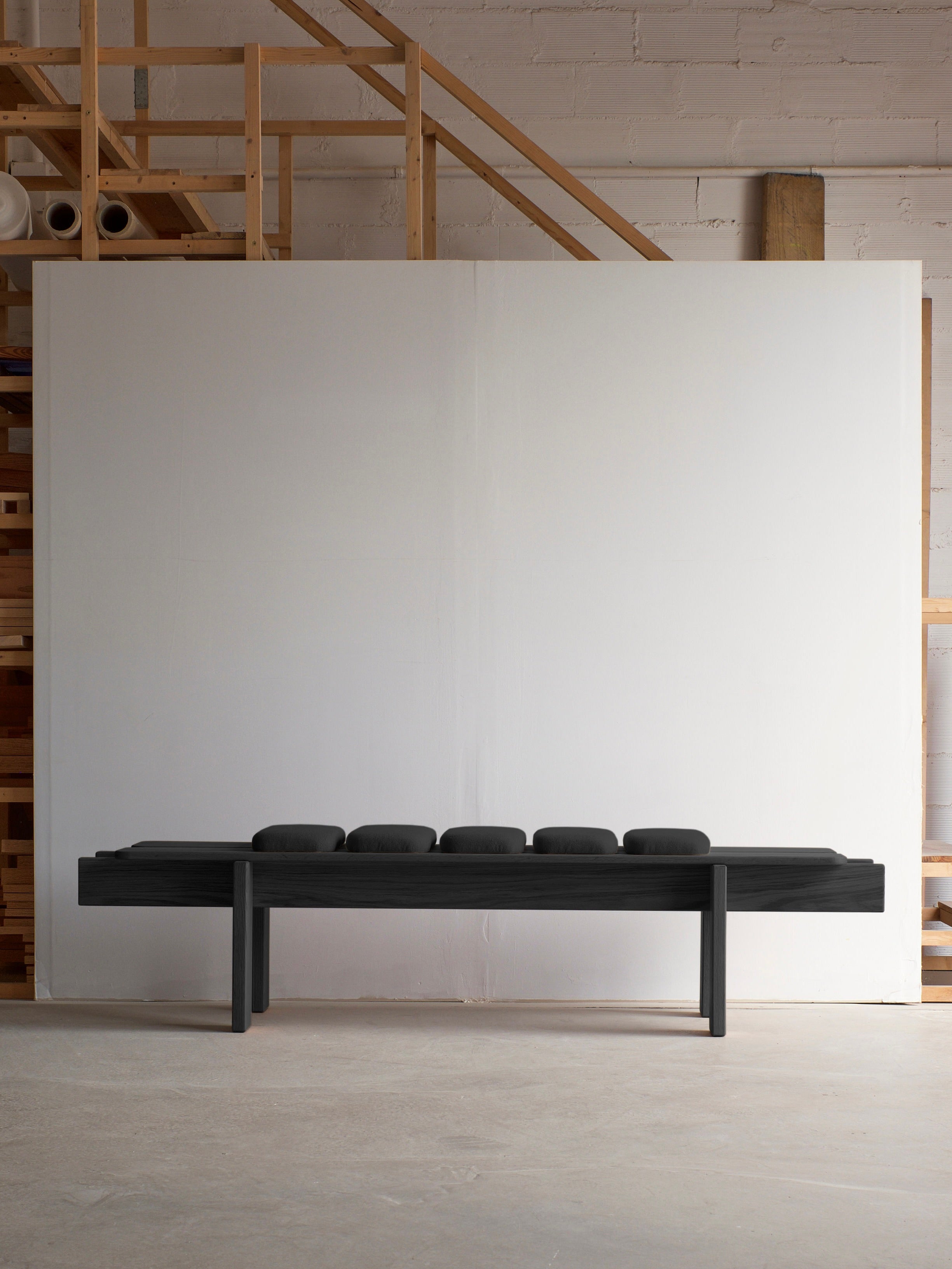 The Ueno Bench Black Edition by malva, featuring five circular cushions and Japanese craftsmanship, is placed in front of a large blank canvas and wooden structures in an industrial studio with concrete flooring and brick walls.