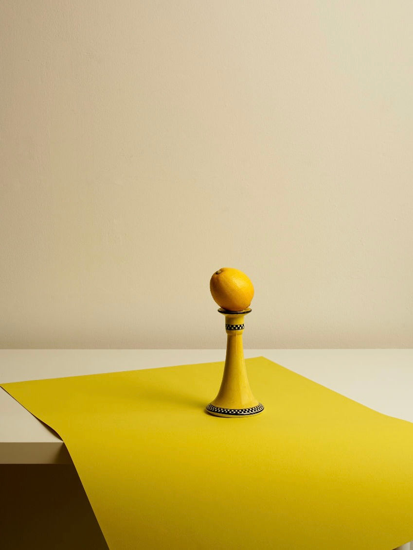 A single yellow lemon is balanced on top of the Porcelain Candle Holder 1920's by Les Objets Amsterdam, showcasing its black and white Art Deco design. The setup rests on a light beige surface, partially covered by a yellow sheet. This elegant arrangement evokes the charm of Czechoslovakia in the 1920s, with a plain beige wall serving as the backdrop.