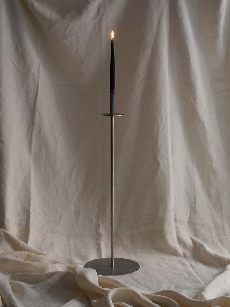 A tall black candle on the FEBRERO SHOP Fusta Candle Holder, crafted from brushed stainless steel, complements the draped, light fabric background for a minimalist elegant setting.