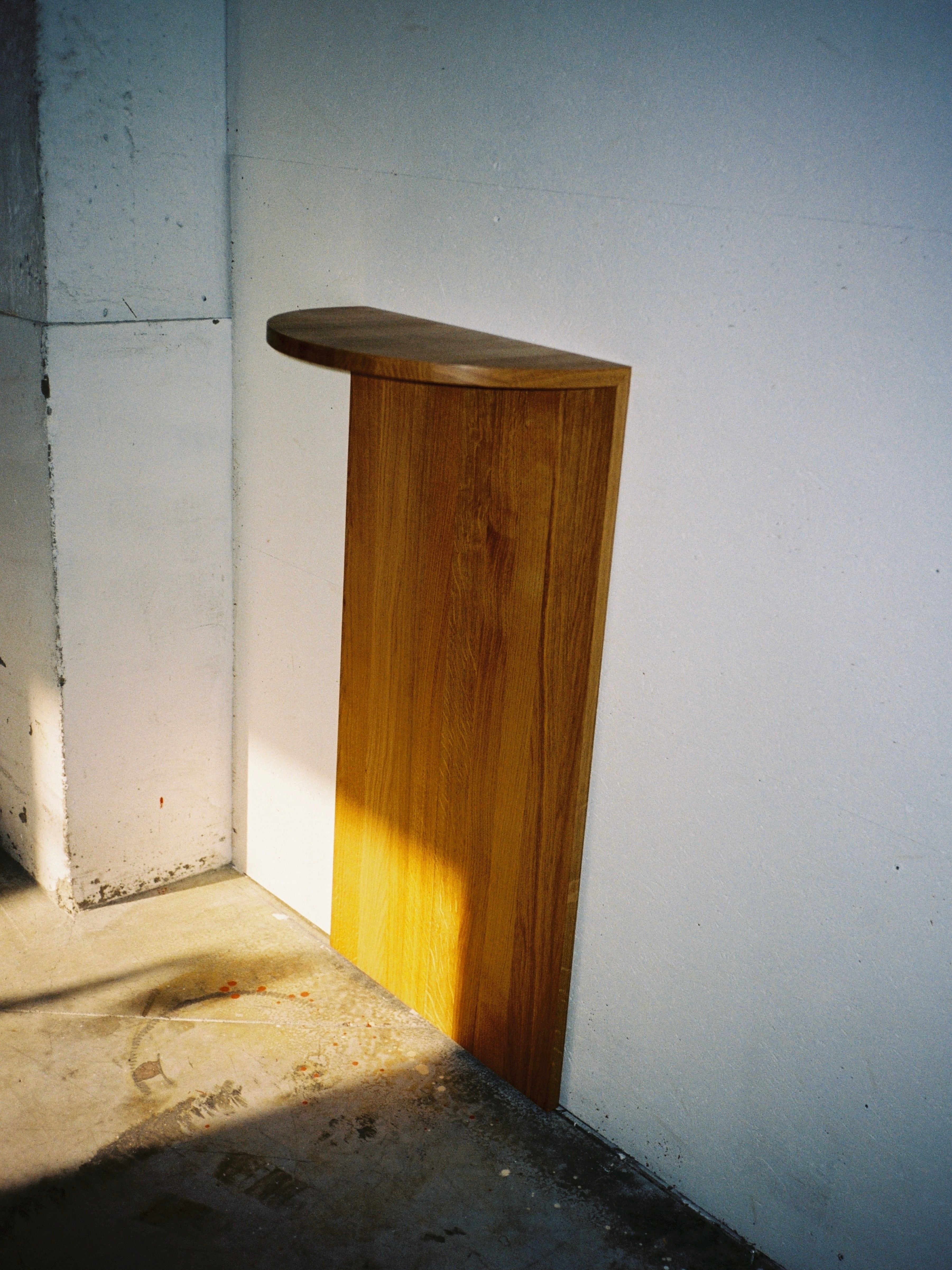 A Megaron Shelf (Large) by COSEINCORSO, crafted in wood and made in Italy, is attached to a white wall in a minimalist setting. The shelf casts a shadow on the ground and wall, illuminated by sunlight from the left. The floor is concrete with some marks and stains.