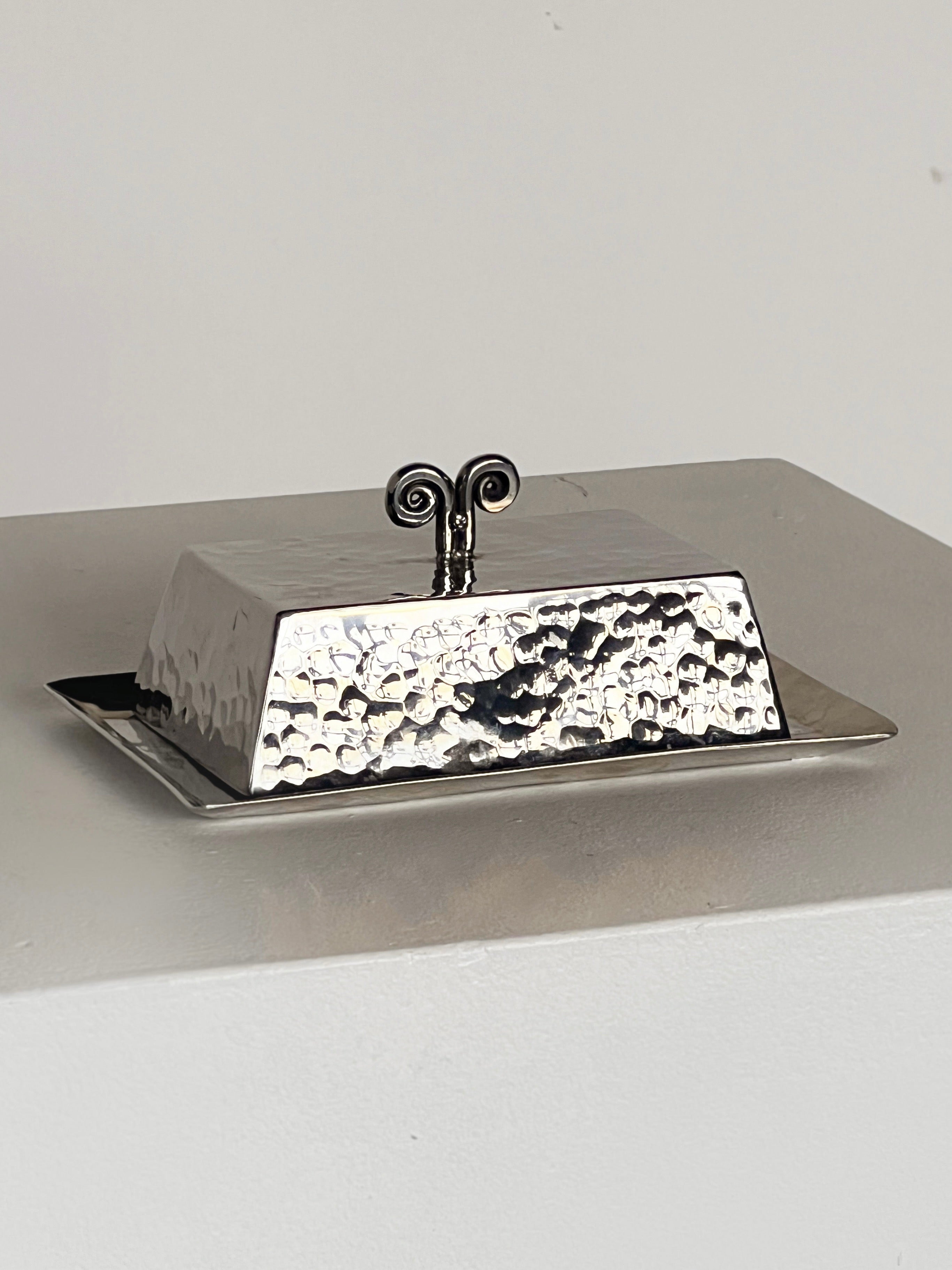 The Spiral Butter Dish by Porter Studio, with a hammered rectangular design and decorative handle, sits on a light surface. The lid exudes 1970s charm, featuring a handle with two outward-facing swirls.