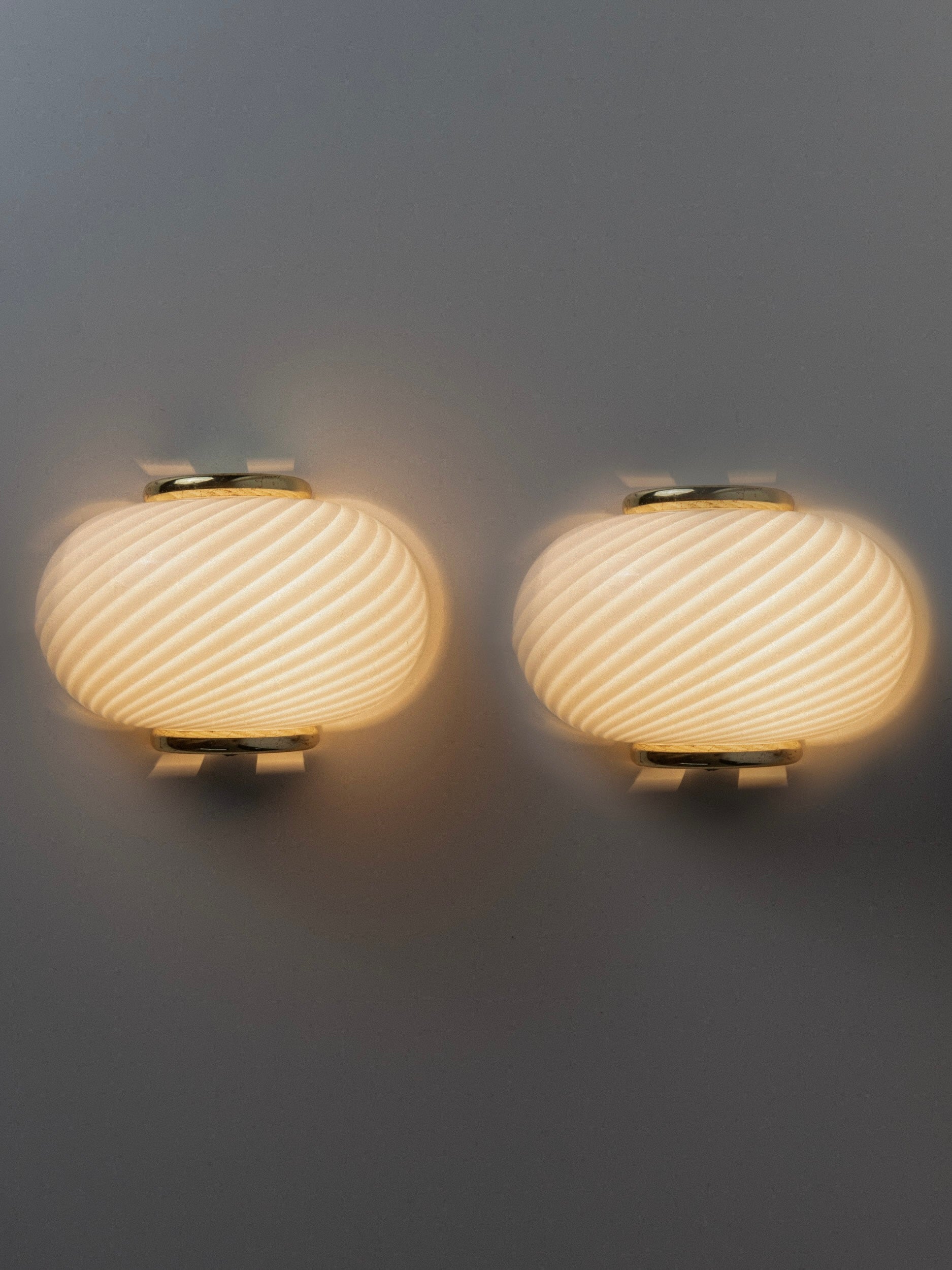 Bottega Jacobs' pair of Murano Glass Wall Lamps offers a modern twist on vintage style, featuring spherical shapes and a textured swirl pattern that softly lights up grey walls with a warm glow, enhancing the elegant design.