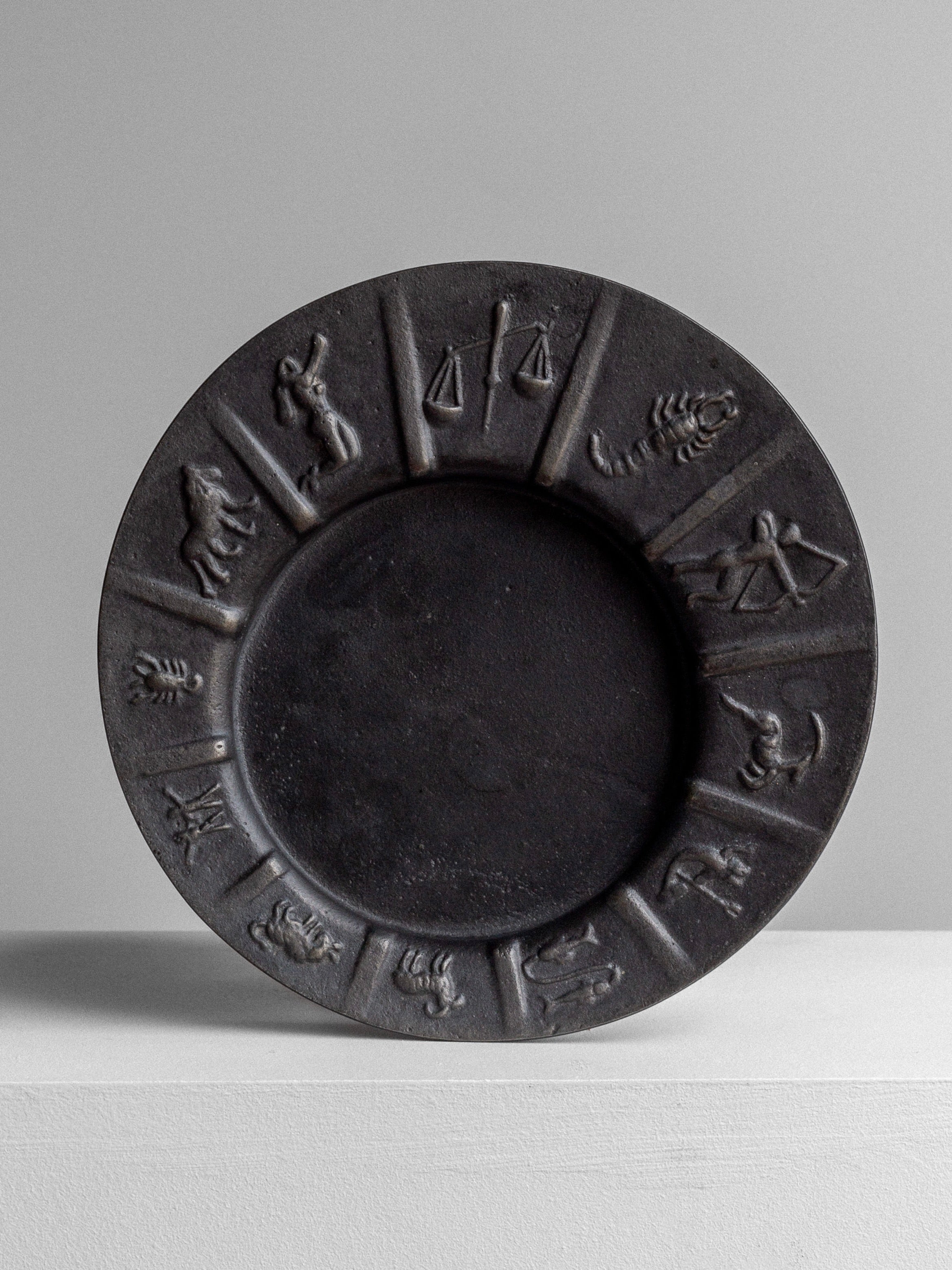 This vintage Cast Iron Zodiac Dish from Spigel is a round, black plate showcasing an intricate zodiac motif with embossed signs, such as scales and a scorpion. It is displayed upright on a white surface against a grey background.