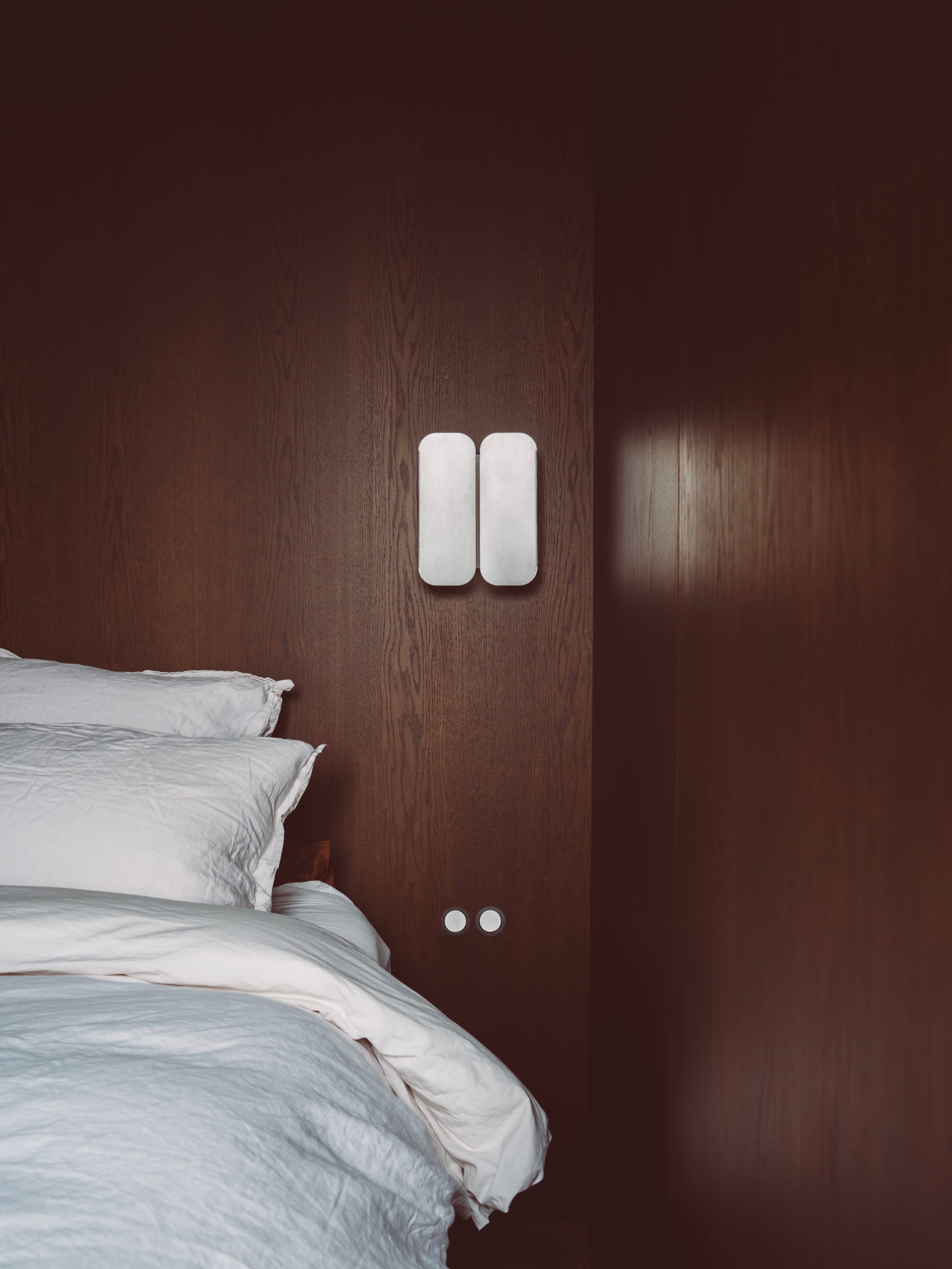A neatly made bed with white linens sits against a dark wood-paneled wall. Above, the KØGE Design Wall Lamp 01 adds to the room’s minimalist, cozy vibe and modern style with its stainless steel, dual-panel design.