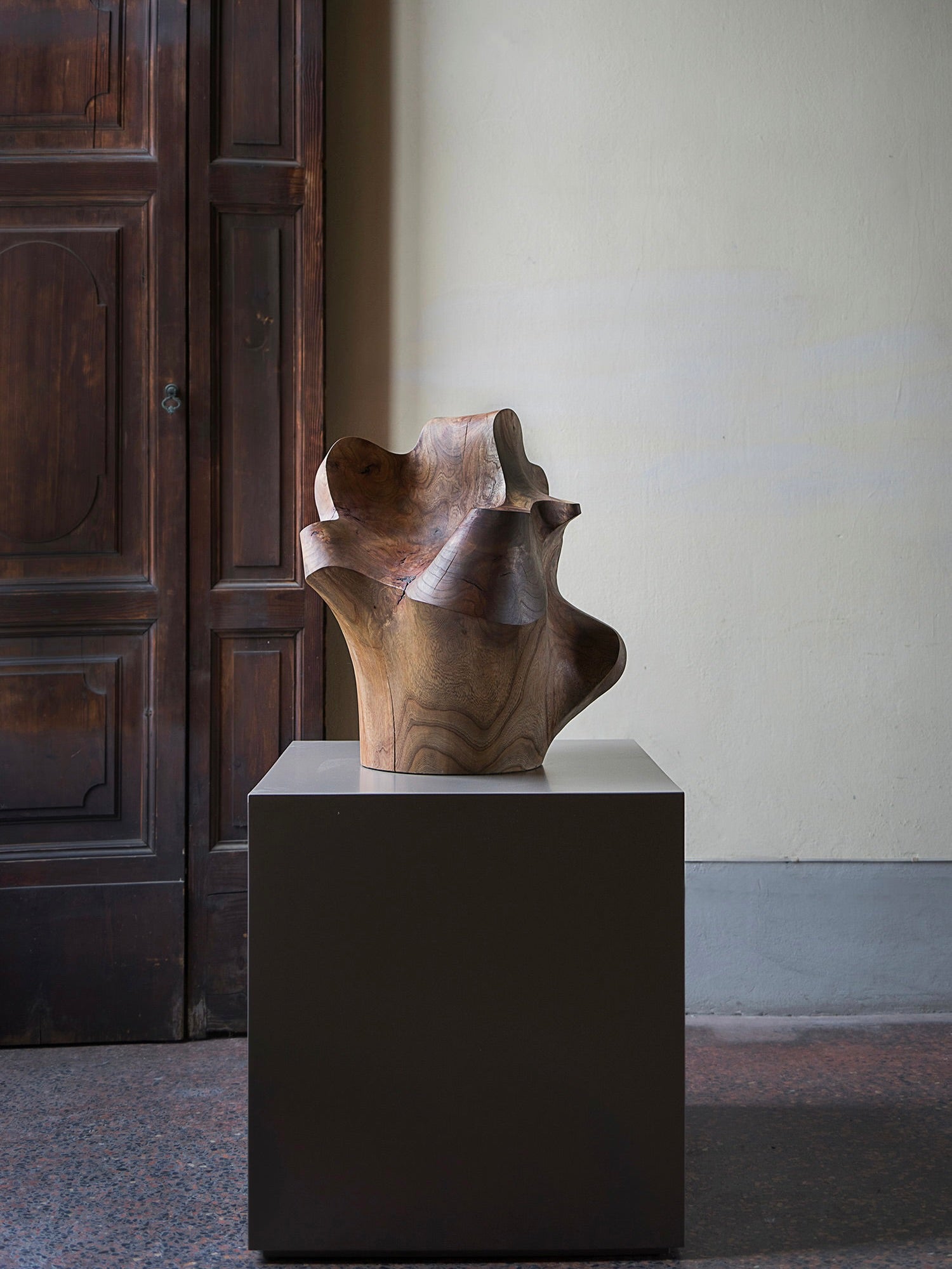 The Sculpture "The Root" by Touch With Eyes is a smooth, handcrafted piece with flowing curves on a dark square pedestal. A rustic wooden door and pale yellow wall in the background highlight the contrast between its organic form and architectural elements.
