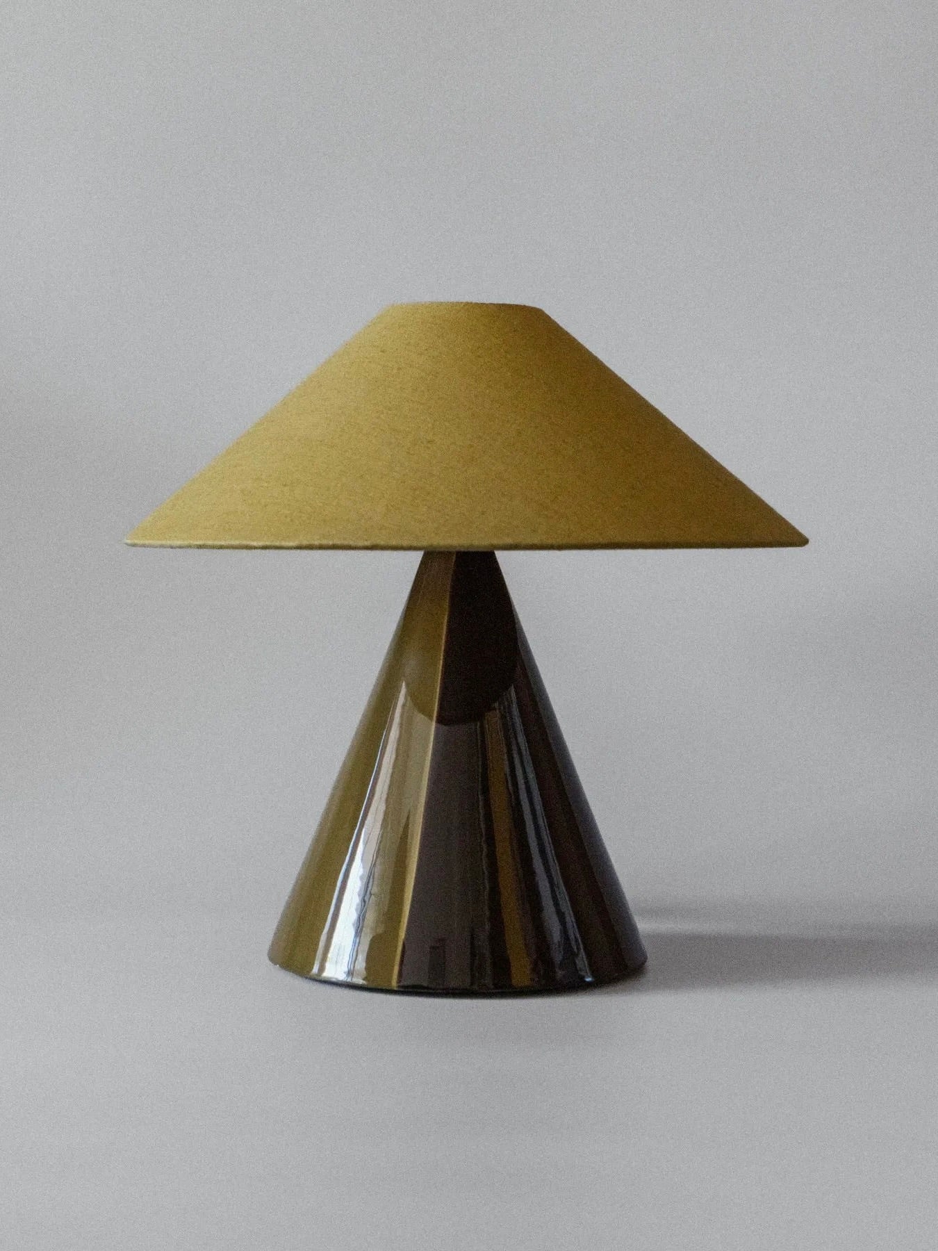 The "Caterina" Golden Olive Table Lamp by mattina moderna is a modern limited edition piece with a conical design, featuring a shiny brown base akin to earthenware and a matte yellow shade, elegantly set against a plain light grey backdrop.