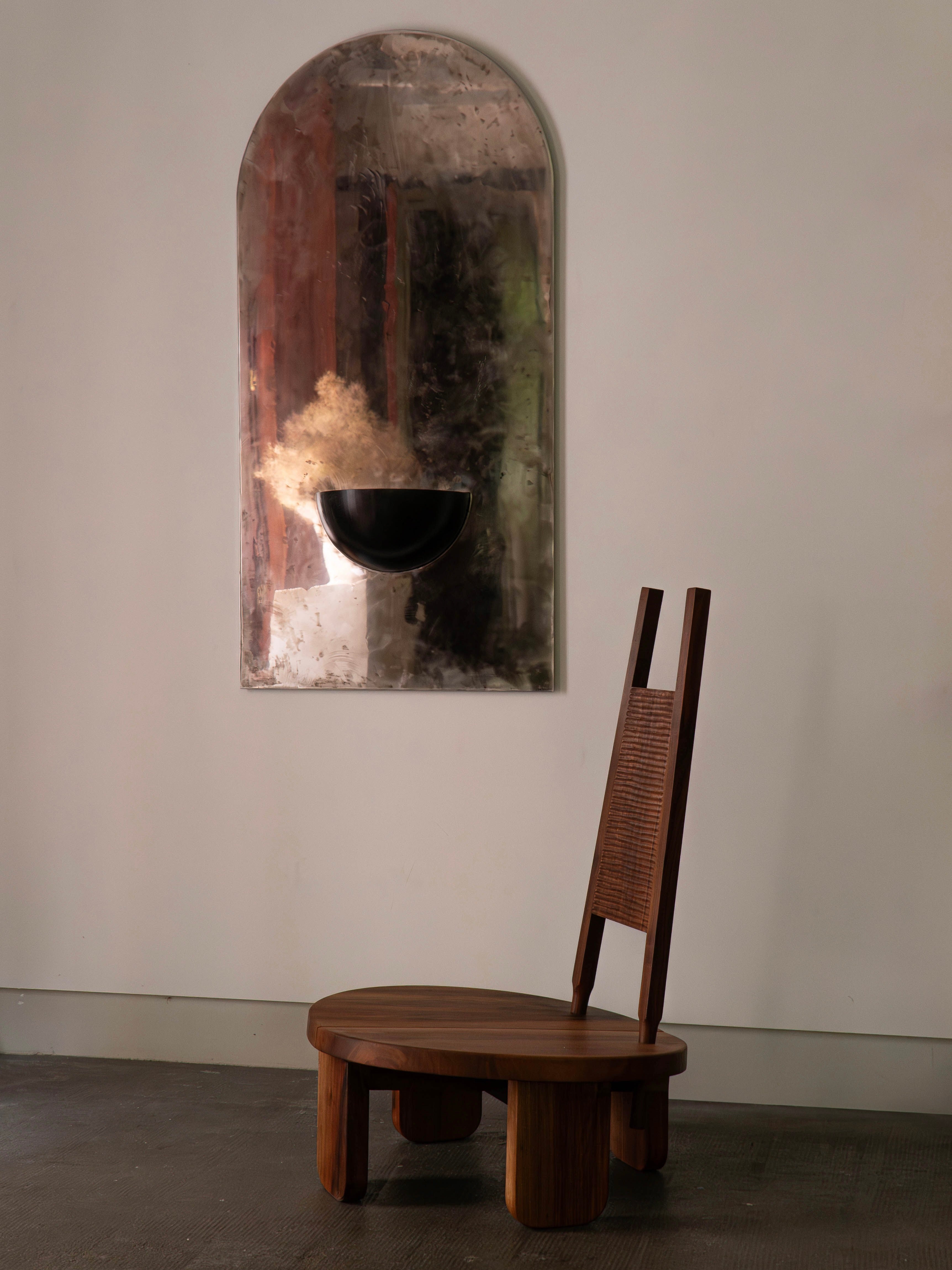A wooden chair with a high backrest is positioned against the wall, above which COSEINCORSO's large Mirror of Simple Souls displays a black bowl filled with dried flowers. The scene maintains a minimalist and artistic vibe, capturing spiritual serenity with its warm and muted tones.