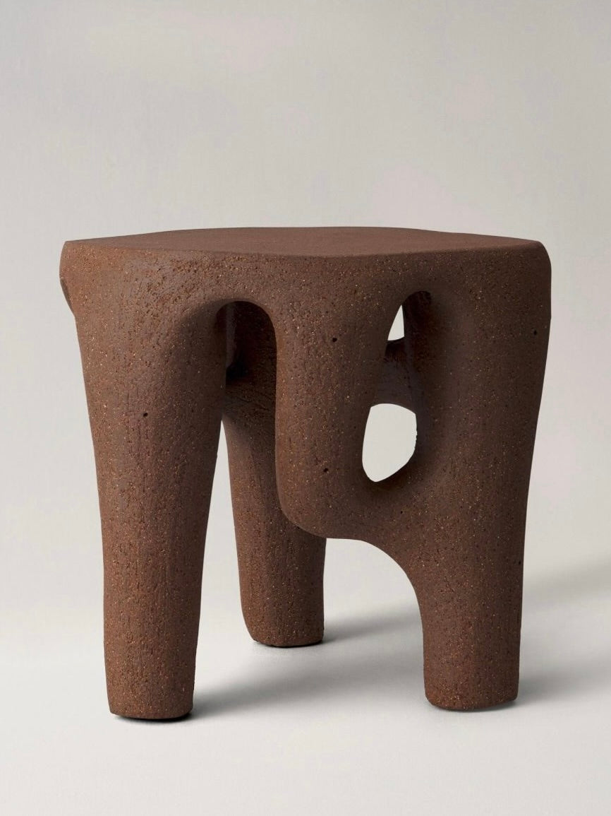 Introducing the Satellite Gatekeeper Stool Sculpture by Abid Javed, a distinctively shaped, abstract creation made from textured brown material. This modern, sculptural design showcases three legs—one adorned with an open oval space—and a slightly irregular top surface that evokes the appearance of stoneware ceramic.