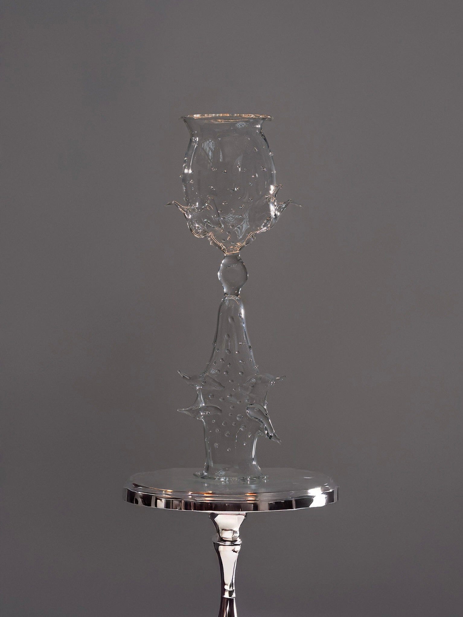 The Objects of Affection_Besos by Pia Glassworks is a delicate, intricately designed glass sculpture resembling a goblet atop a shiny metallic stand against a gray background, featuring ornate, abstract details that exude an enchanting aura.