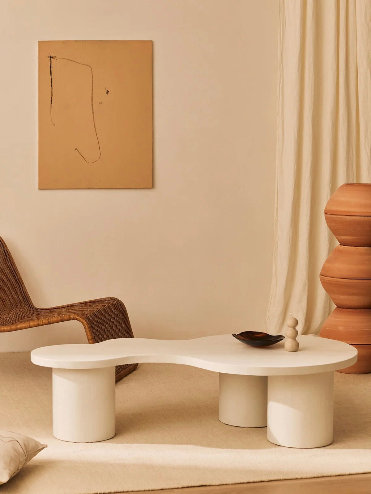 A minimalist room showcases a MarlotBaus Ahumado Coffee Table with organic curves on a beige carpet, alongside a wicker chair, geometric wooden sculpture, and simple abstract painting. Cream curtains complement the space's serene atmosphere.