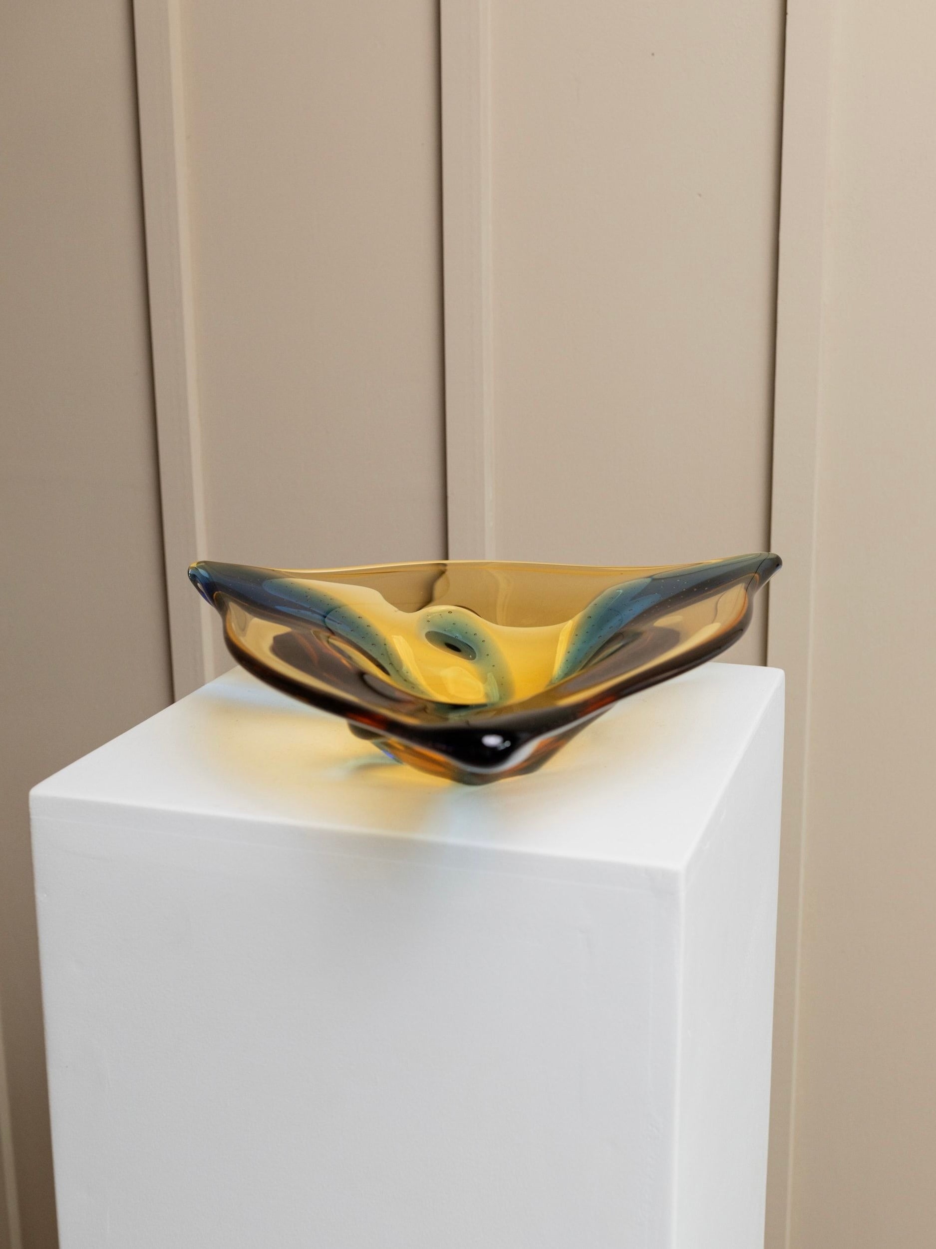A Vintage Glass Bowl by Karel Zemek, featuring swirling amber and teal hues reminiscent of vintage glassworks Mstisov, is elegantly displayed on a white pedestal. Crafted by Bottega Jacobs, the bowl's edges curve gracefully, capturing and reflecting light. It is set against a backdrop of beige paneled walls.
