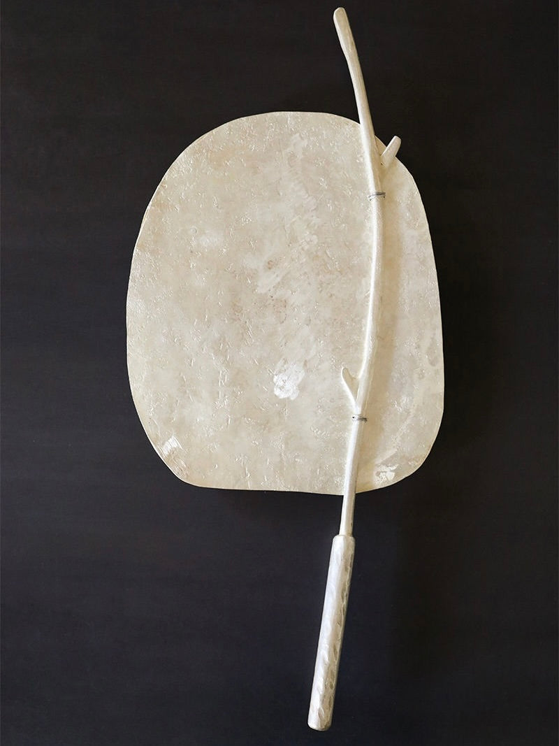 A modern sculpture from Rooms Studio in Tbilisi, named the Silver Plated Casted Cake Stand, features a large, irregularly-shaped beige stone with a textured surface mounted on a dark background. Attached to it is a slender, curved, branch-like element in a lighter, whitish hue, creating an elegant contrast between the two shapes.