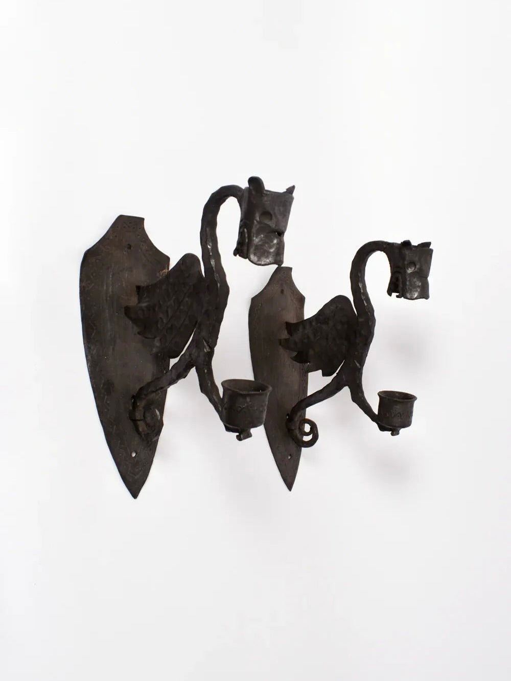 A pair of Barracuda Interiors mural candleholders from the 1960s, crafted in France, featuring medieval-style zoomorphic dragons with detailed wings and curled tails. Each dragon holds a candle holder in its mouth, and the dark textured metal is mounted on shield-like backplates, providing a vintage gothic look against a light background.