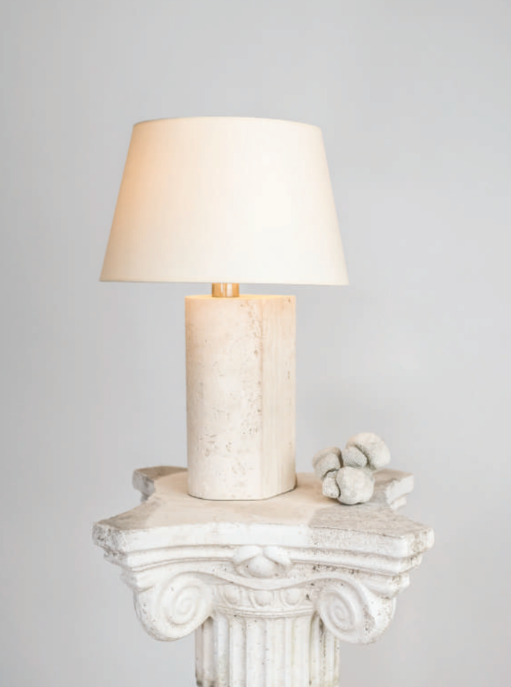 A Capri Travertine table lamp 2 by Brajak Vitberg, with a cylindrical base, sitting on an ornate, classical-style white pedestal decorated with a cluster of stones.