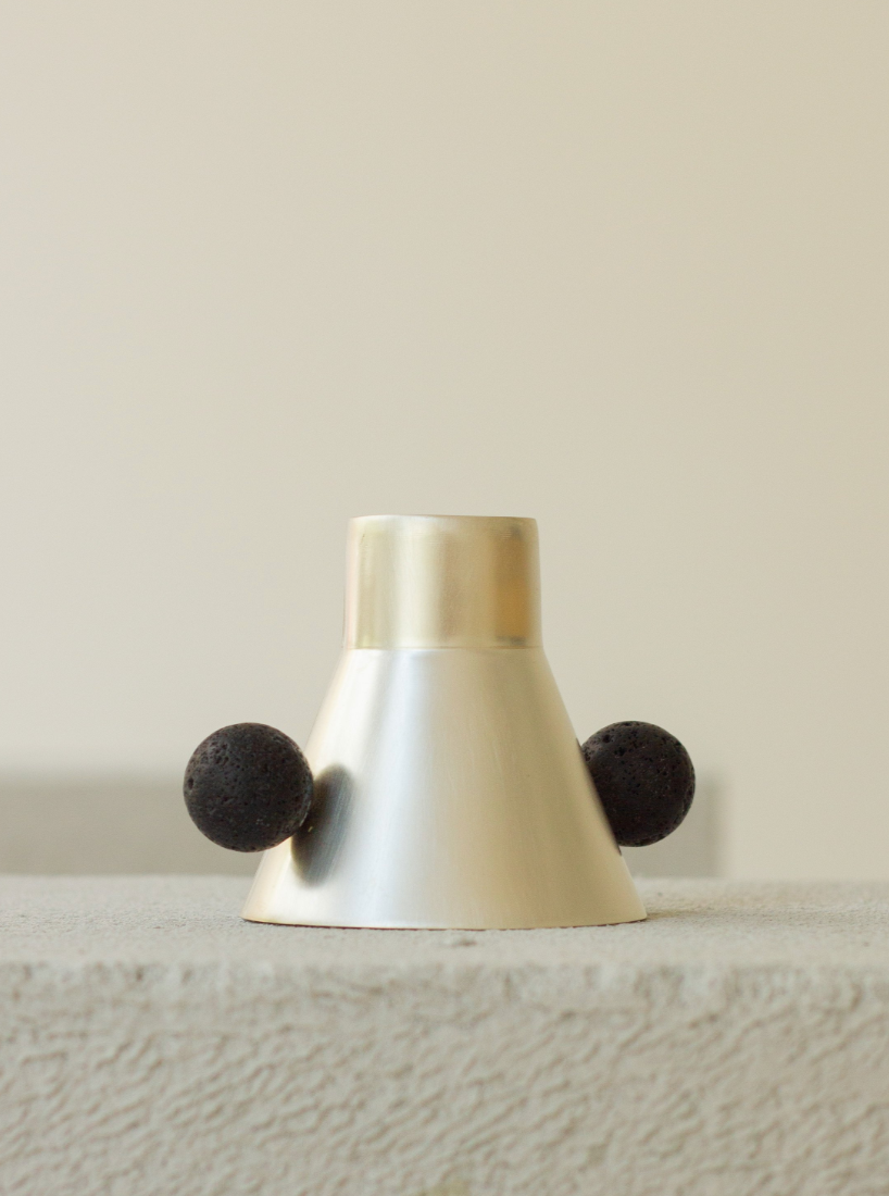 A shiny metallic Candeliere with a single dark, rough-textured sphere attached near its base, positioned on a light gray surface against a neutral background.