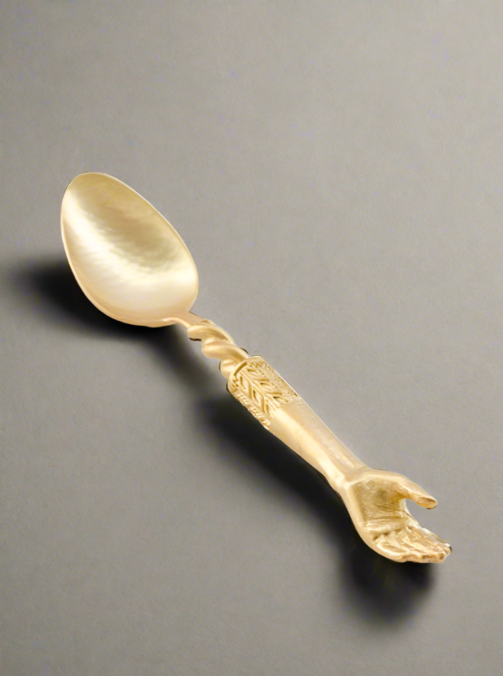 The Hand Tea Spoon by Natalia Criado is a handcrafted golden spoon featuring a unique handle shaped like an upright human hand. Its artistic design adds contemporary flair, making it ideal for luxury interior decor against a plain gray background.