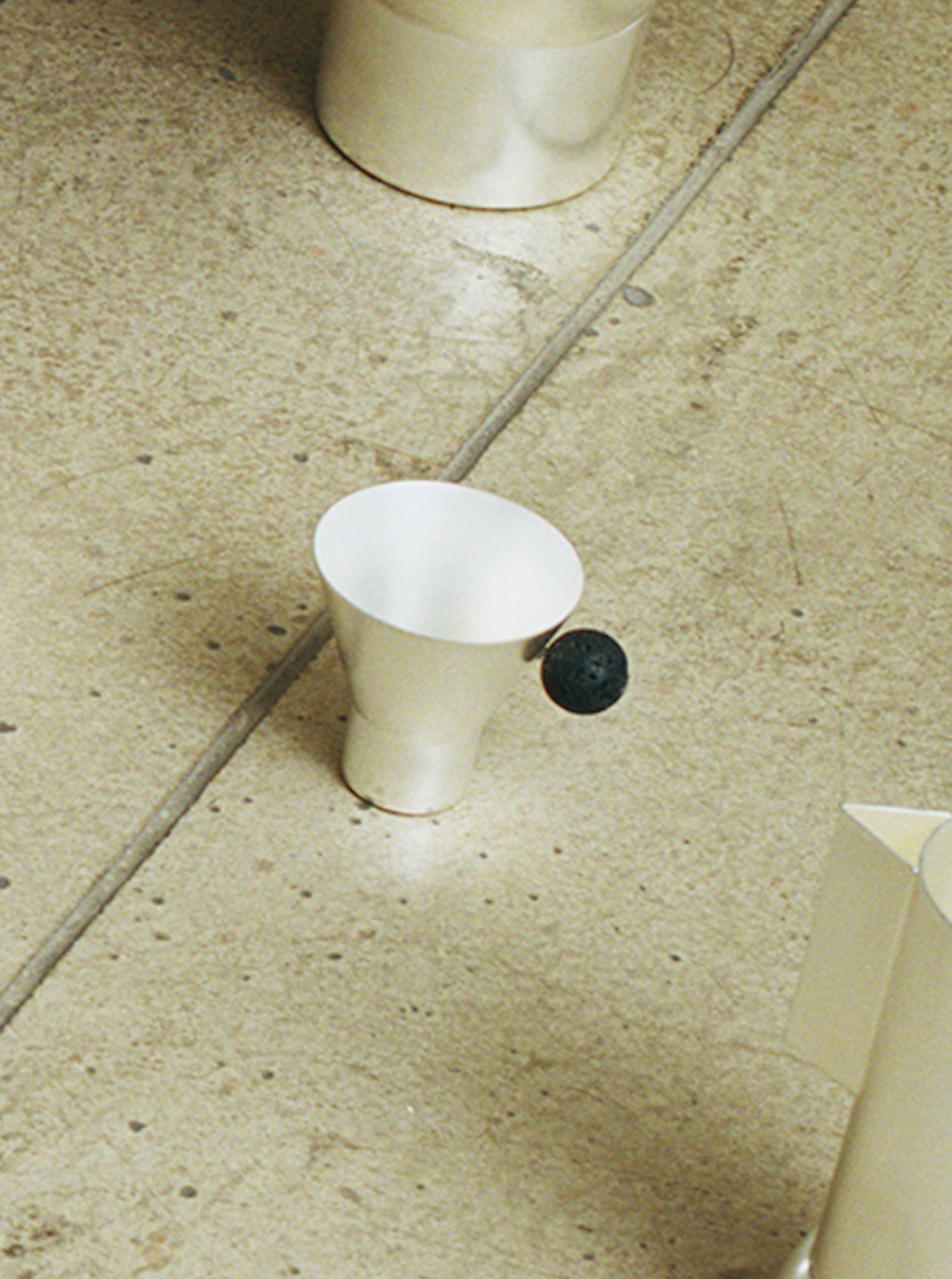 A white paper Cone Cup by Natalia Criado lying on a textured gray tile floor beside a silver-plated metal cup.