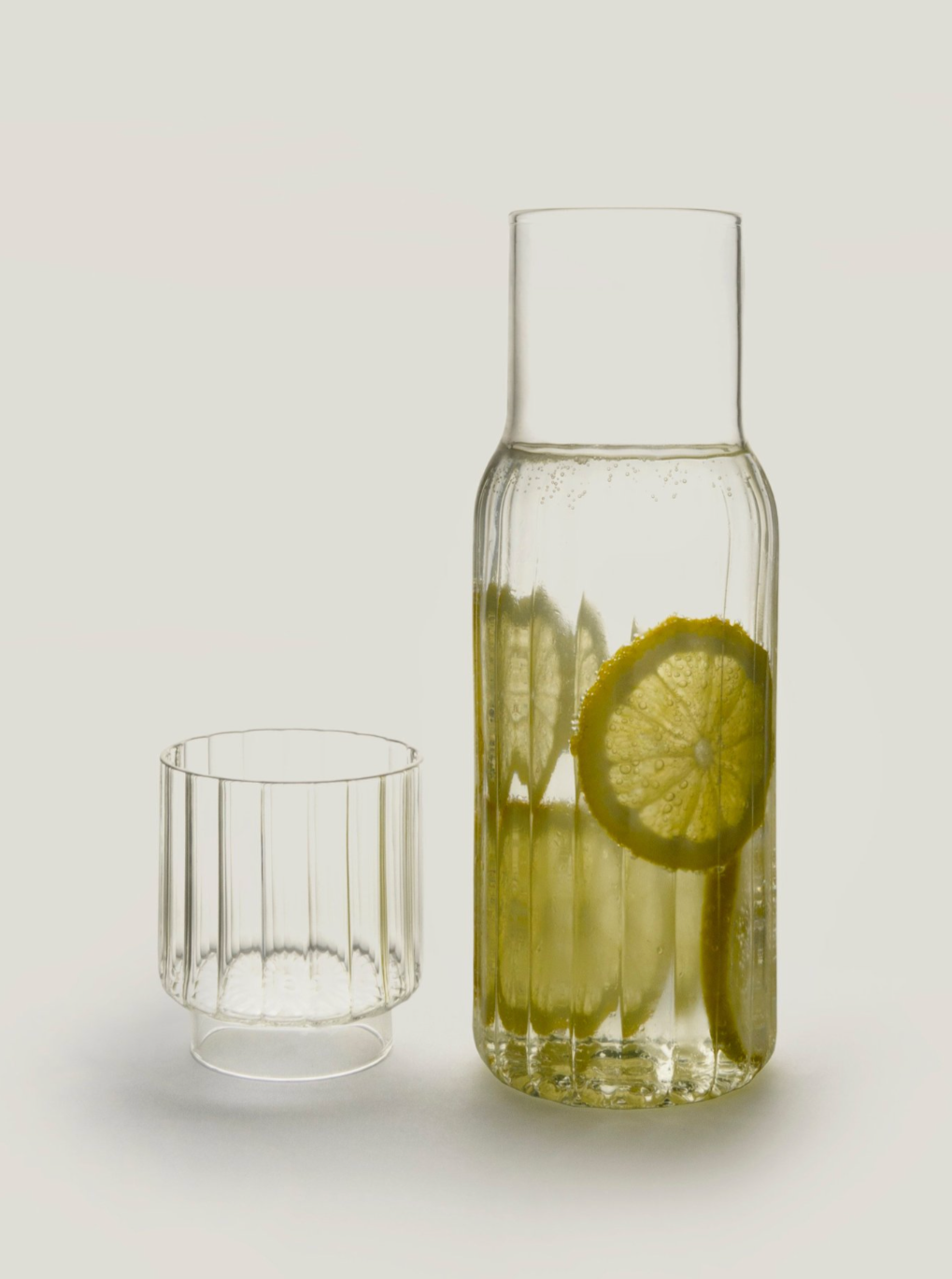 A handcrafted in Italy, Agustina Bottoni borosilicate glass Pillar Carafe Set containing water with a slice of lemon inside, next to a small empty glass on a plain white background.