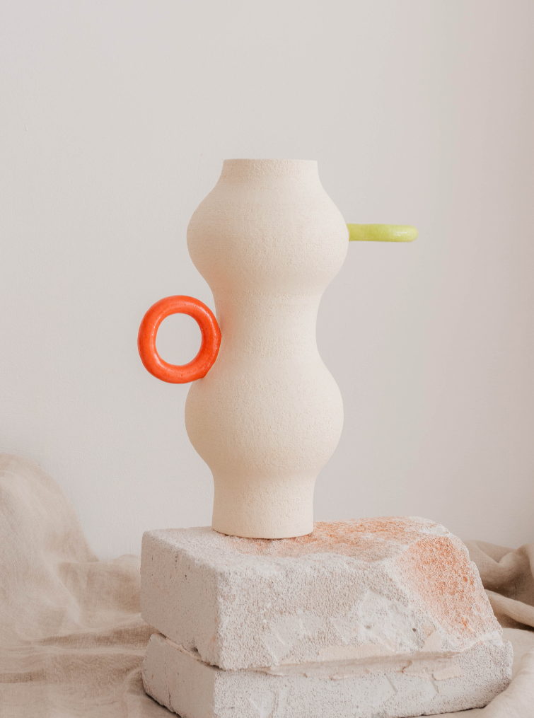 A modern art sculpture featuring an abstract, curvy figure in white grogged stoneware, with a green handle emerging from its side and a red circular object looped through it, standing on Pot-au-feu's VASE 01.