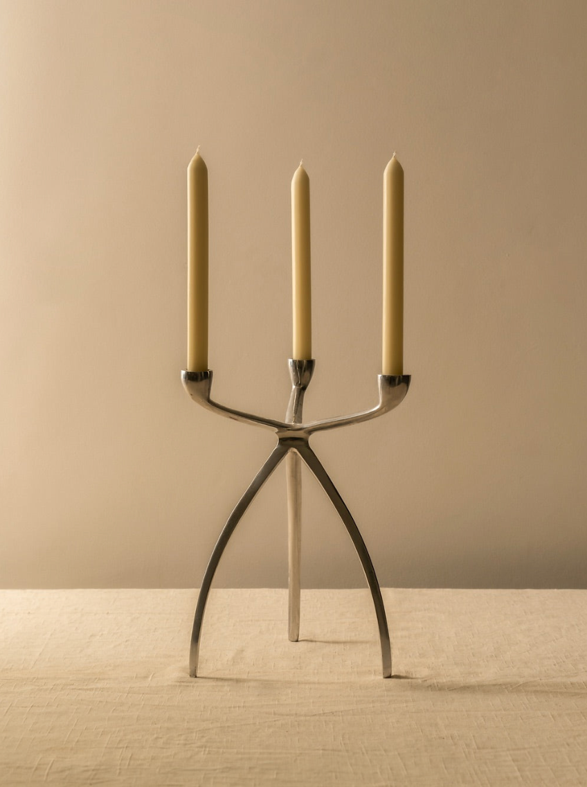 A modern, silver three-armed Tripod Candle Holder by Les Objets Amsterdam stands on a beige table with three unlit, slender, cream-colored candles. The design is minimalist and symmetrical, with a light beige background complementing the overall aesthetic. This retro-inspired decor piece adds a touch of elegance to any space.