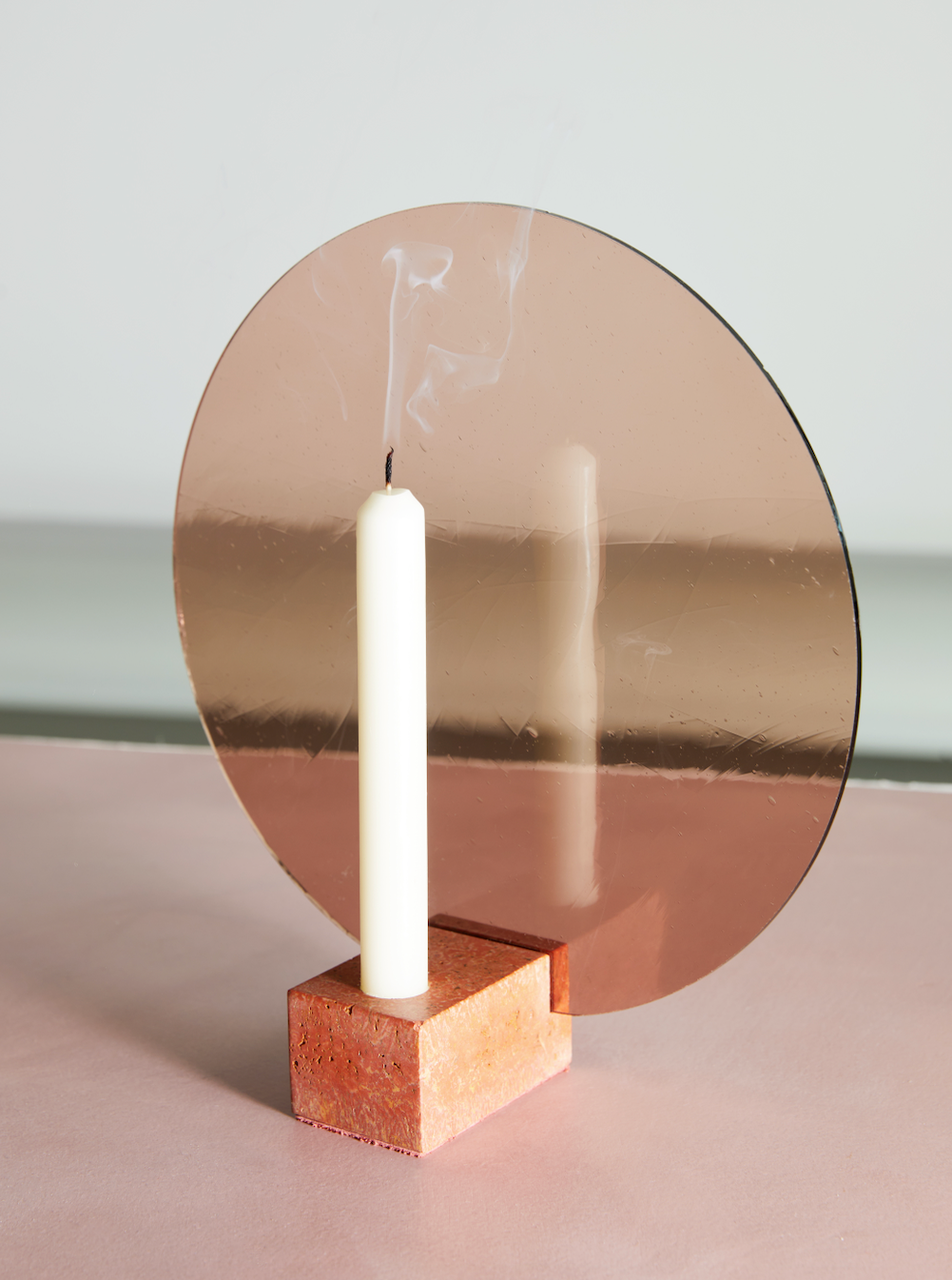 The Aureool - Pink candle by Anto Loua, placed in a handcrafted holder, stands extinguished with smoke curling from the wick. It sits on a square, textured base against a circular, reflective rose-colored backdrop on a soft pink surface against a pale background.