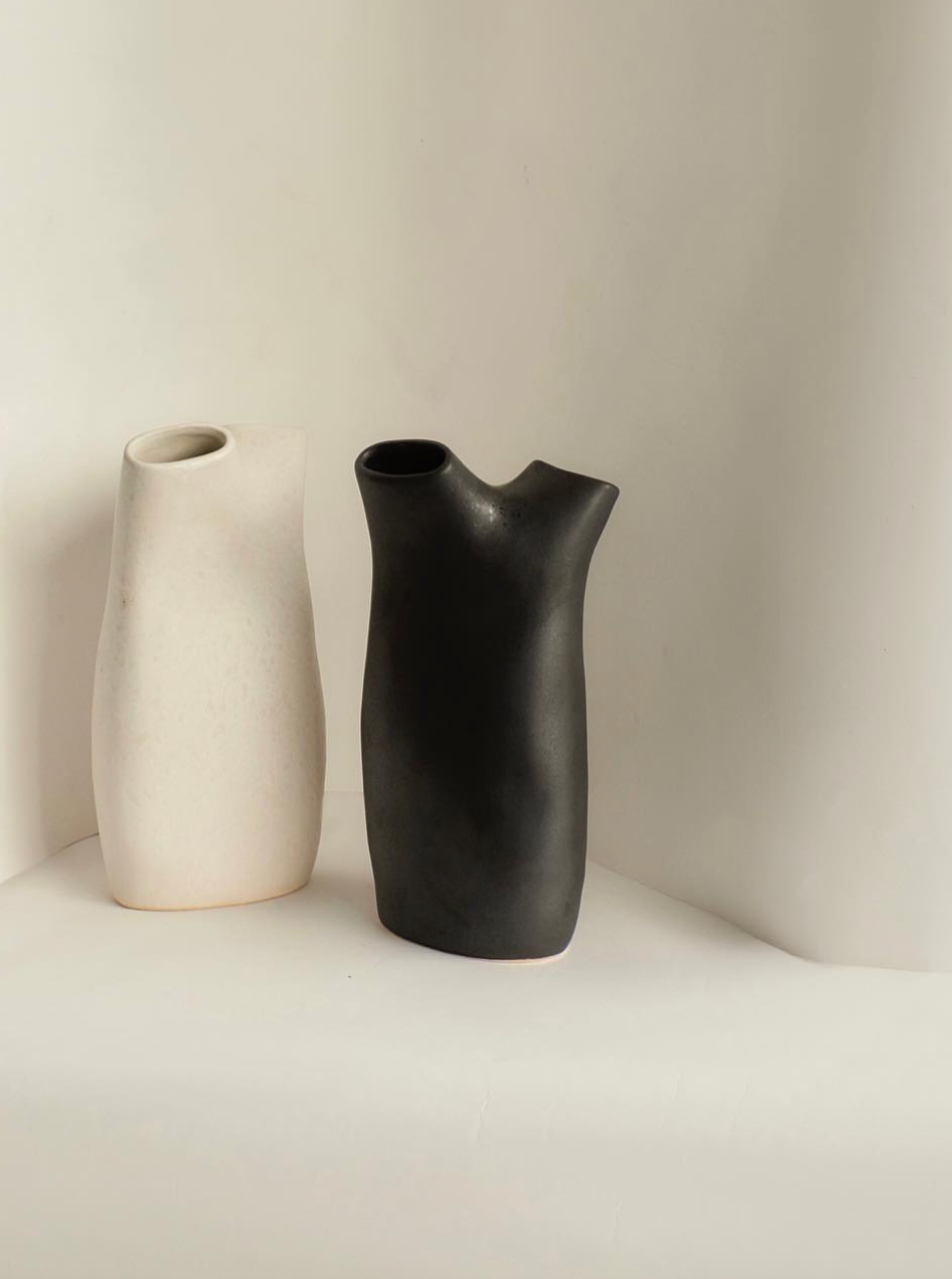 Displayed against a light background are two sculptural ceramic vases by Project 213A. On the left, a white vase features a simple rectangular shape; on the right, the Gemini Vase in Graphite showcases a sleek, contemporary curve. Both vases have small openings at the top.
