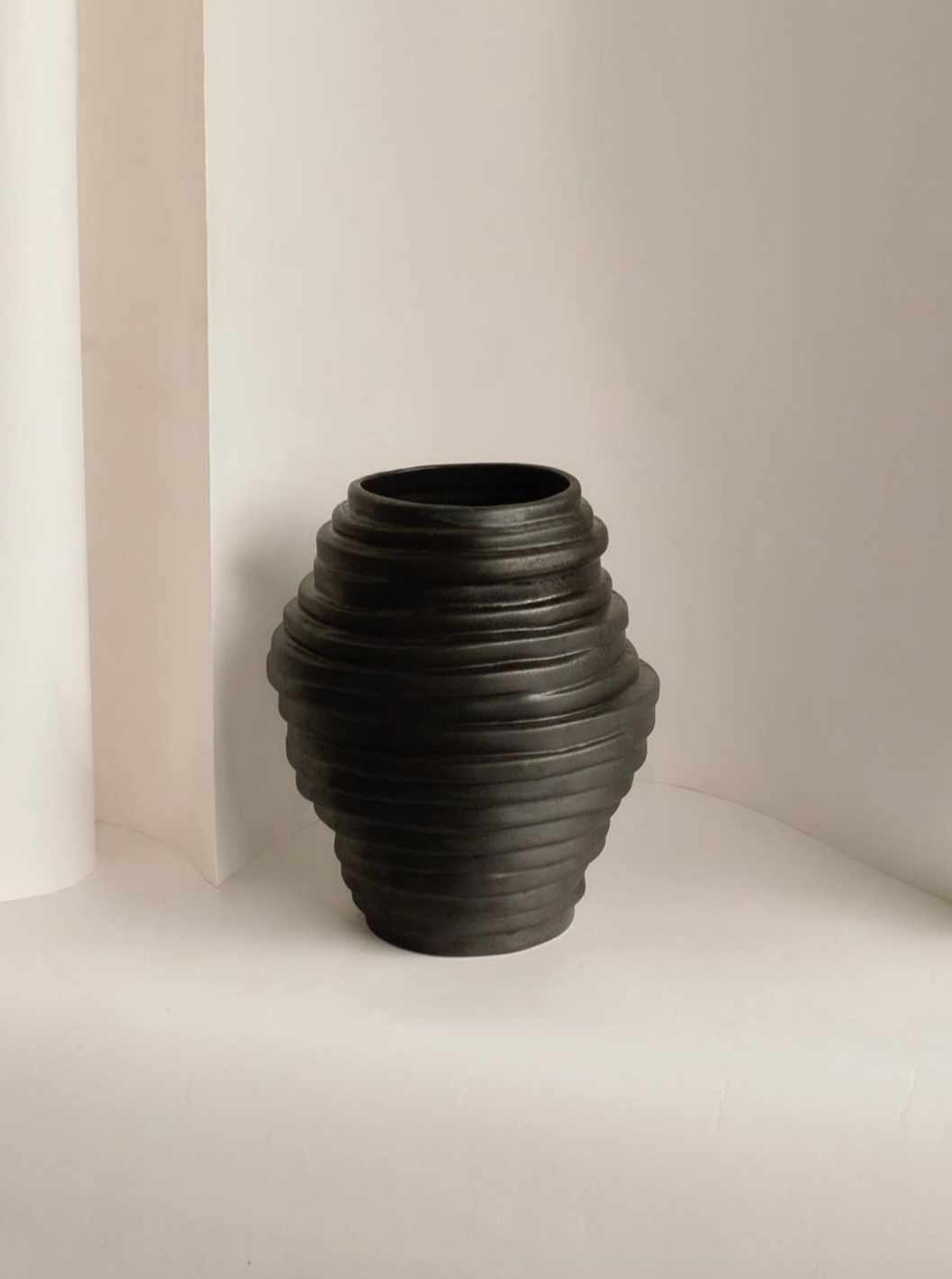 A Alfonso Vase in Graphite by Project 213A with horizontal ridges, displayed against a soft beige background with a simple, minimalistic setup inspired by volcanic rock formations.