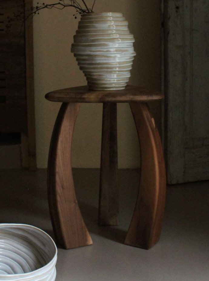 A tall, uniquely shaped white vase with horizontal ridges sits atop an elegant Project 213A stool designed as part of the Arc de Stool '52 collection in a dimly lit room, showcasing a rustic aesthetic.